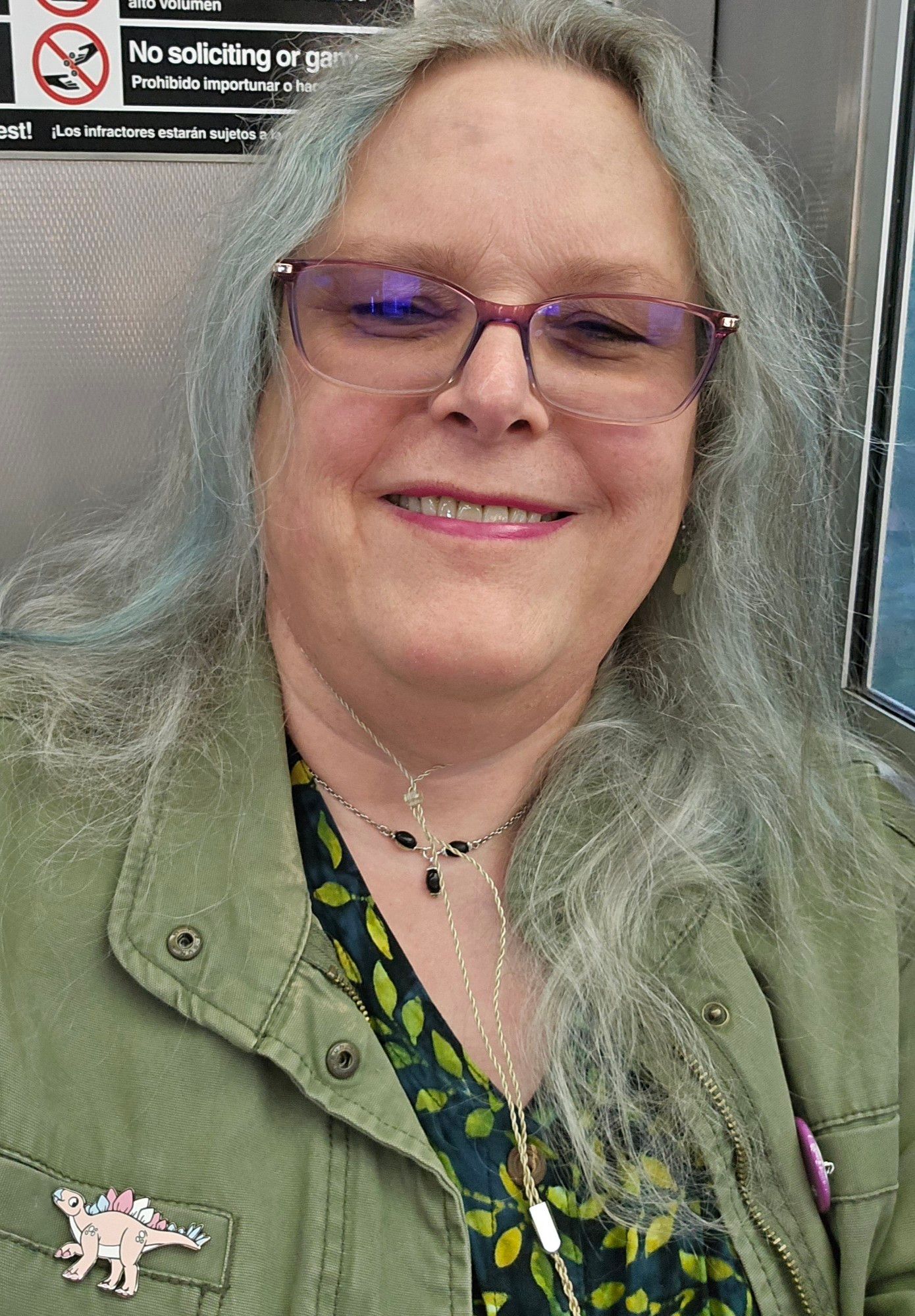 Yeah, another train selfie. Hair down, green denim jacket over dark green print top, trans pride stegosaur pin