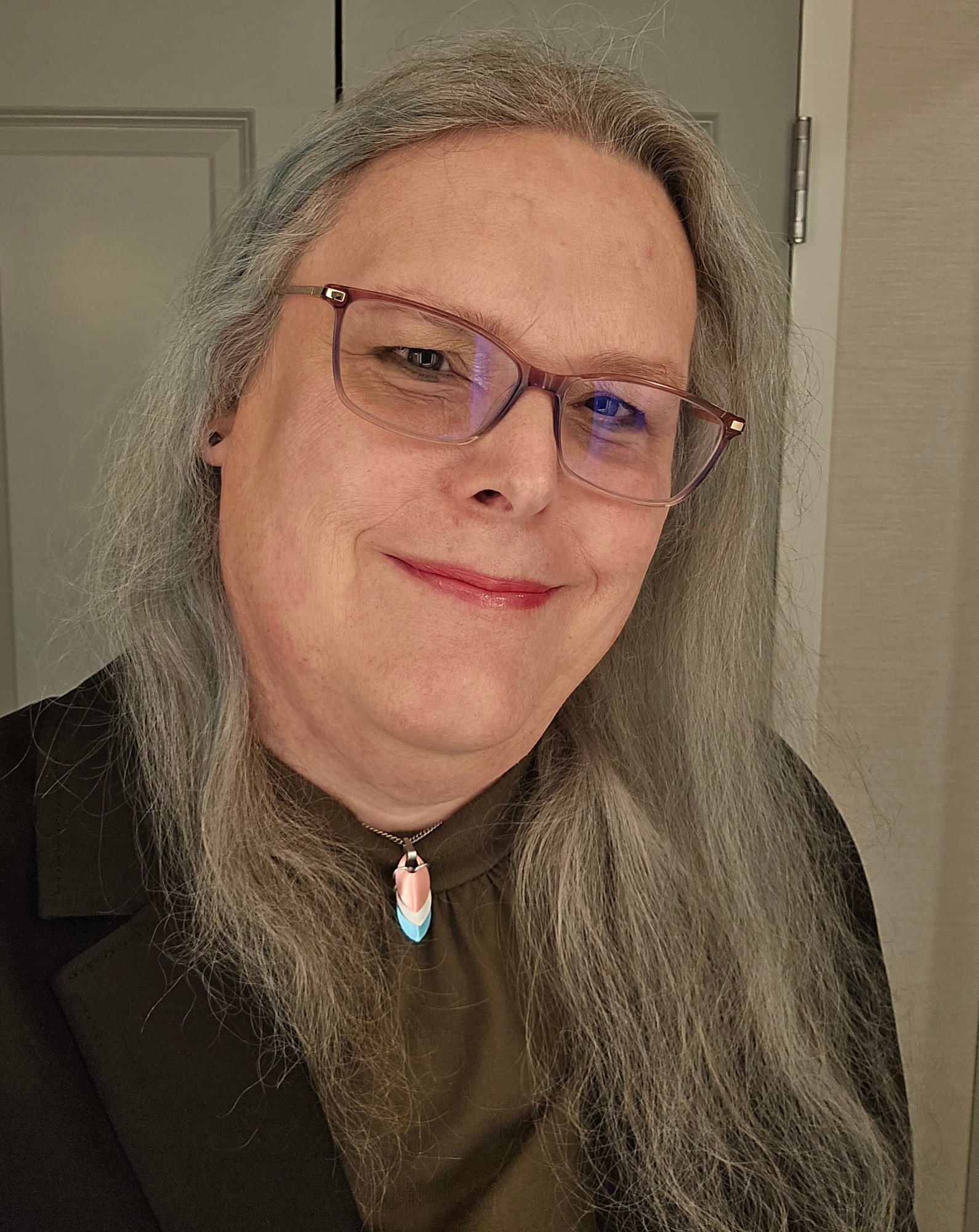 Selfie, hair down, green top, trans pride necklace.