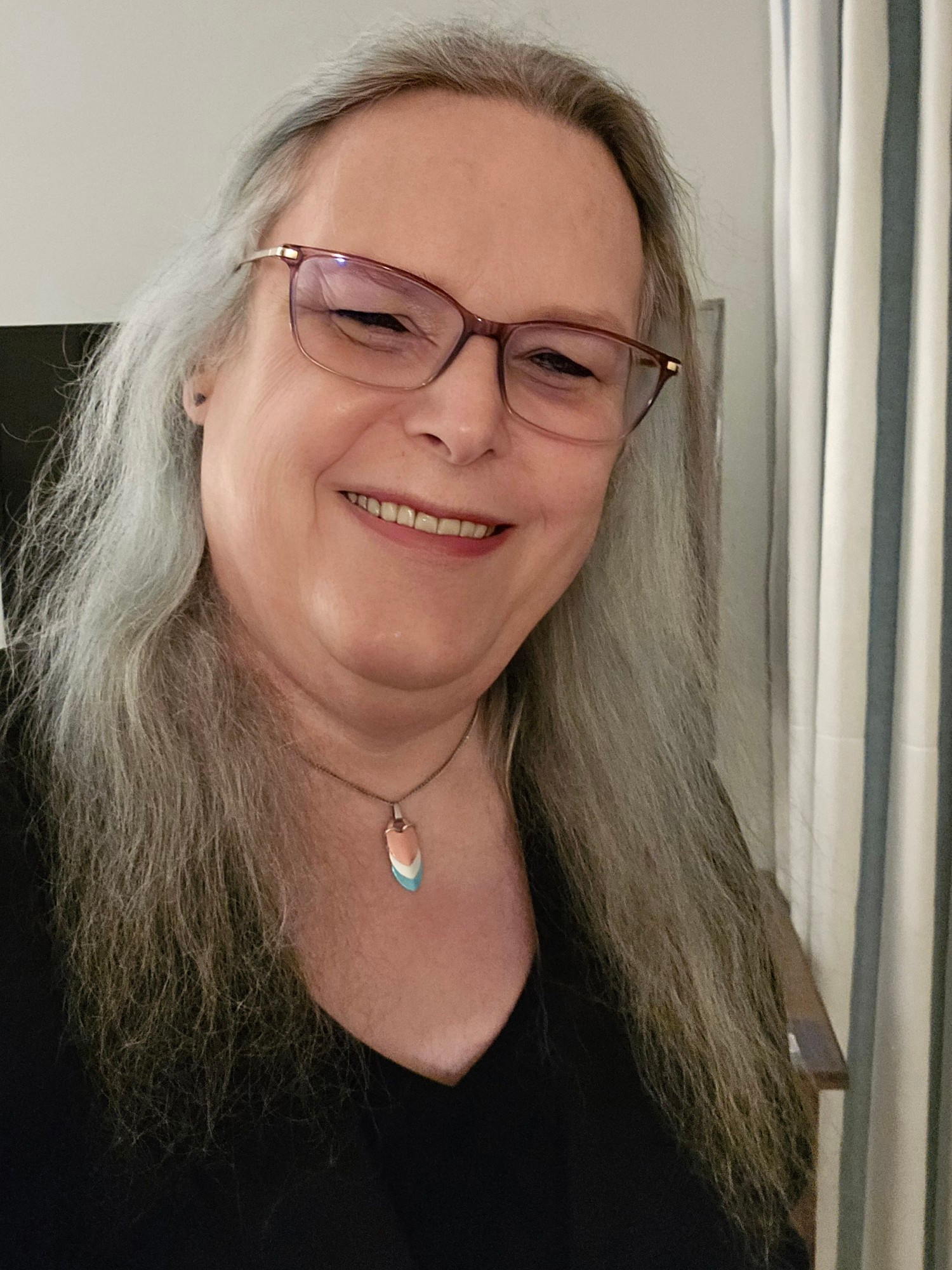 Selfie, hair down, black jacket and top, trans pride necklace.