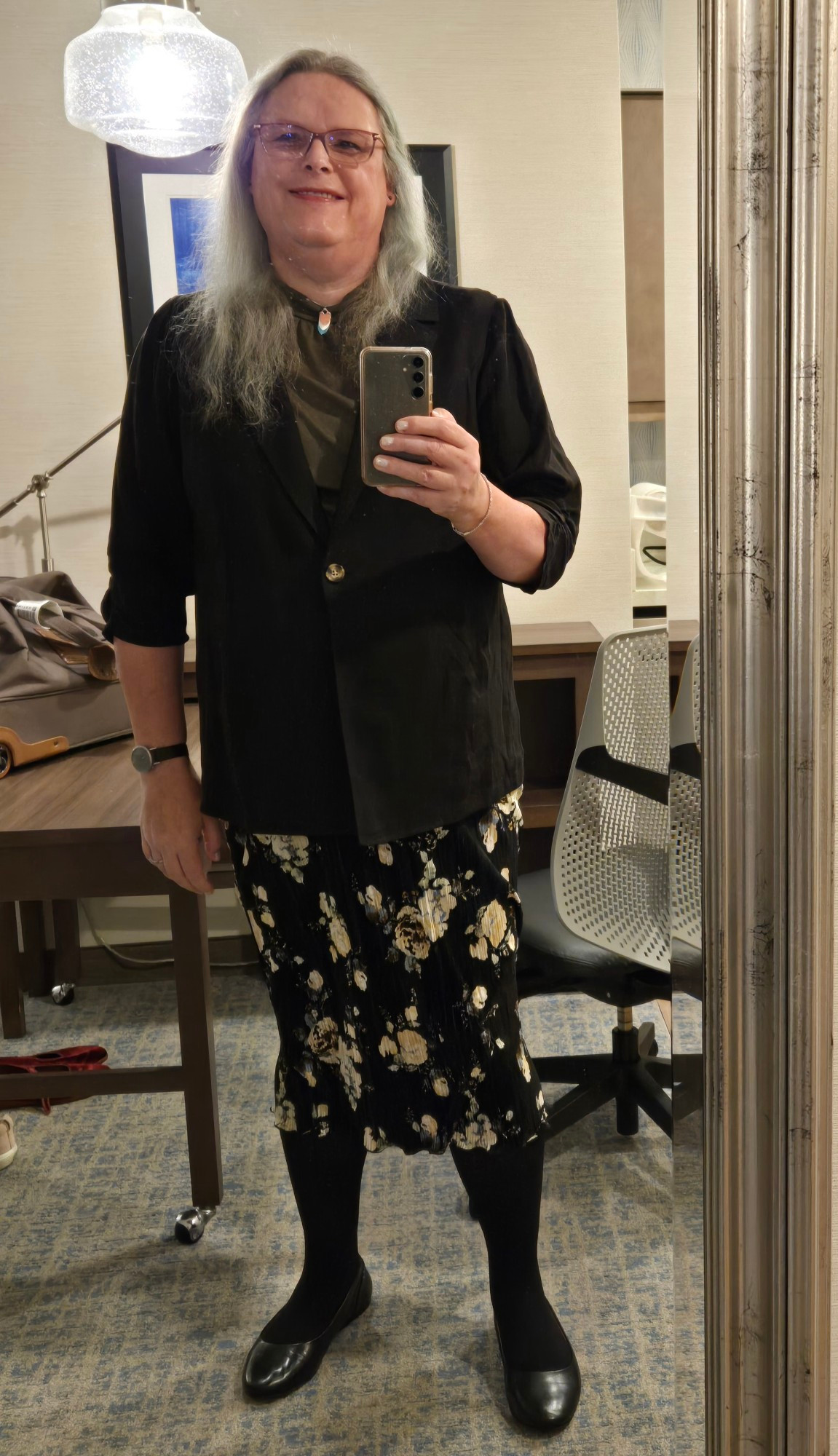 Full-length shot: hair down, black blazer over green top, black calf-length skirt with floral print, black tights and flats.