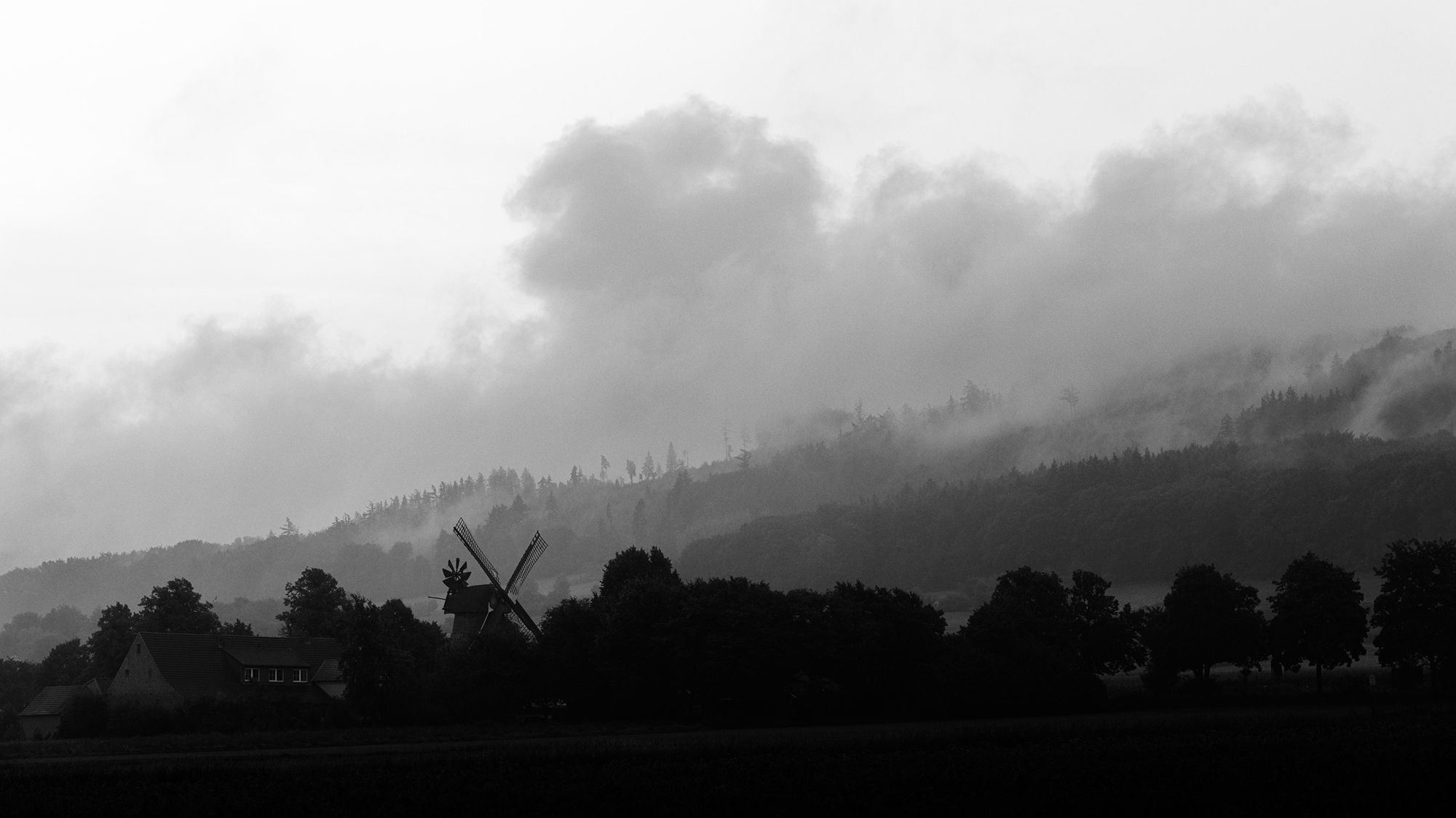 Monochrome photo of Clouds over a hill with a windmill and trees in the foreground