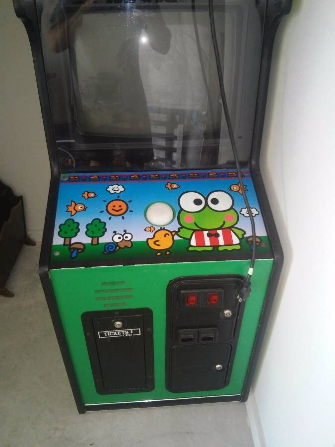 A Keroppi arcade cabinet. It has a big cartoon frog, a yellow duck/chicken, trees, and a sun around a large singular white button. The machine's screen is dark.