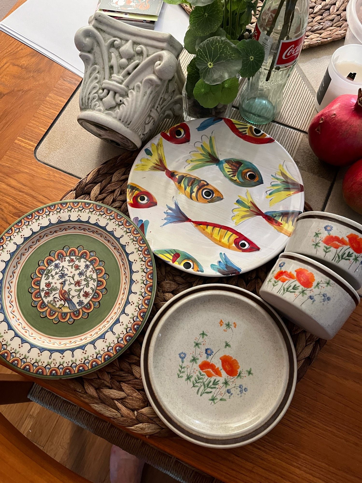 A flat lay photograph of six vintage-looking ceramics. Three are plates with flowers, fish, birds, and other lovely hand painted patterns in shades of red, orange, yellow, deep blue, and green. There are two matching ramekins with a matching orange poppy flower design and also a Grecian pillar flower stand.