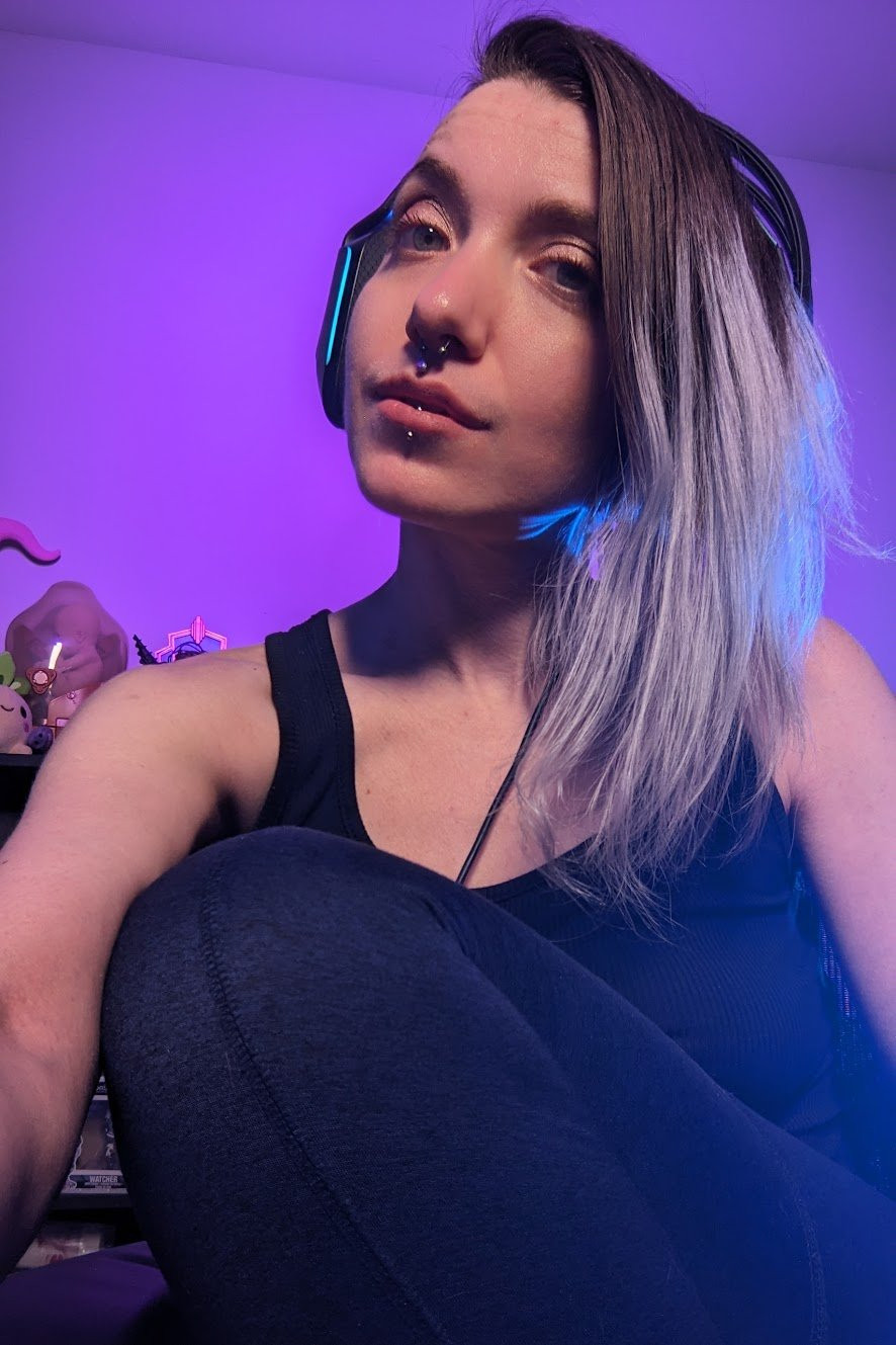 Amaris Skye, a person with light-colored hair is seated in a room, wearing a black tank top and headphones with neon accents. Their hair cascades down to one side, with strands catching a bluish hue from the lighting. The background features a soft purple glow, with various objects like figures or collectibles partially visible, adding some texture to the setting. Their facial expression is calm and relaxed, and they have piercings in their septum, philtrum and lower lip. The ambient lighting creates a distinct and vibrant atmosphere.