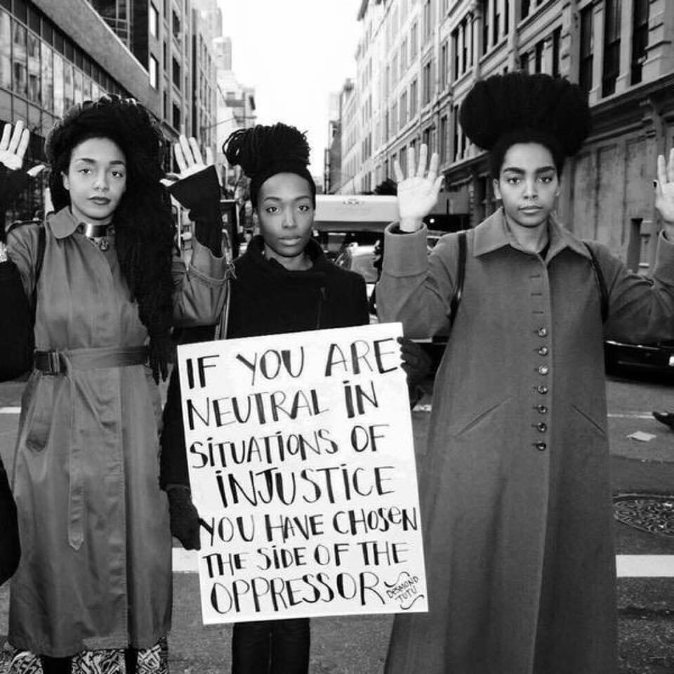 Three black women, two with their hands in the air and the other holding a sign that says “If you are neutral in situations of injustice you have chosen the side of the oppressor” which is a quote by Desmond Tutu