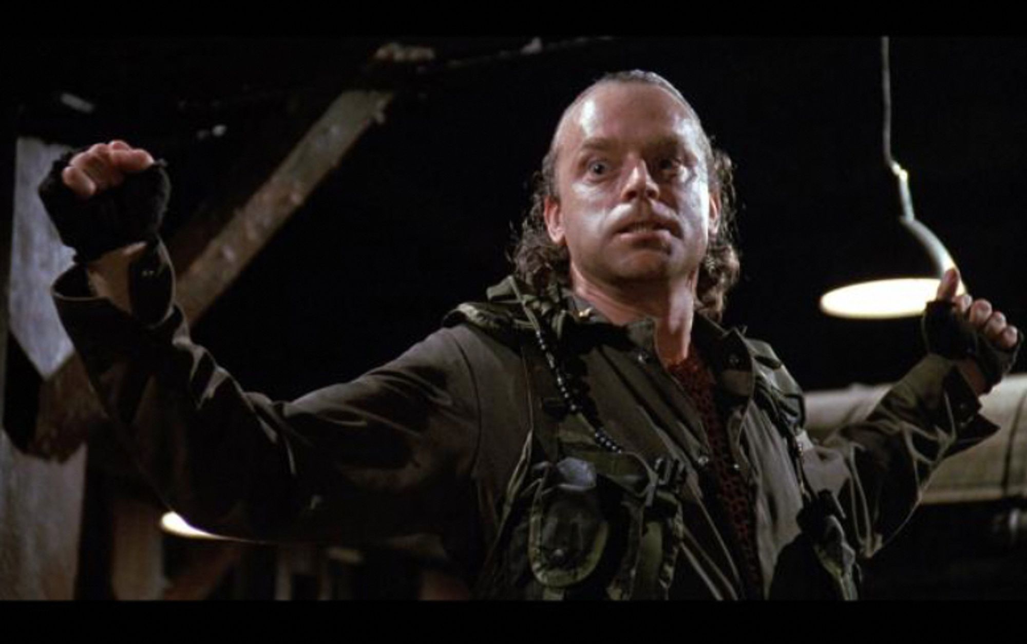 Very unfortunate photo of Brad Dourif's Exterminator character from the 1990 film Graveyard Shift. He's got a receding hairline with hair slicked all the way back, wearing a cargo style boiler suit and cutoff gloves and is palpably sweaty.