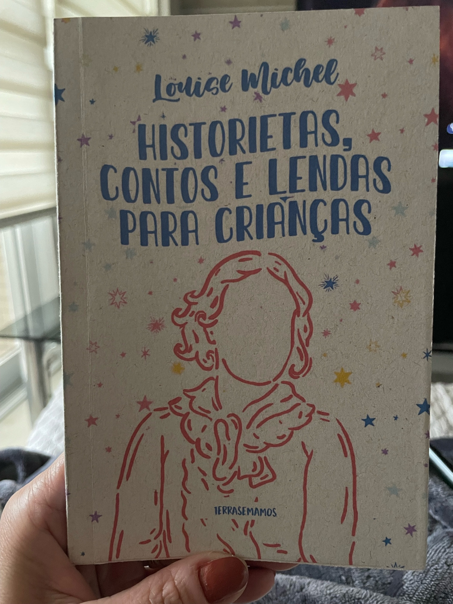 Foto da capa do livro de Louise Michel. Historietas, contos e lendas para crianças da Editora Terra sem anos. Capa de cor clara com estrelas coloridas salpicadas e o contorno de uma figura feminina alusiva a autora.