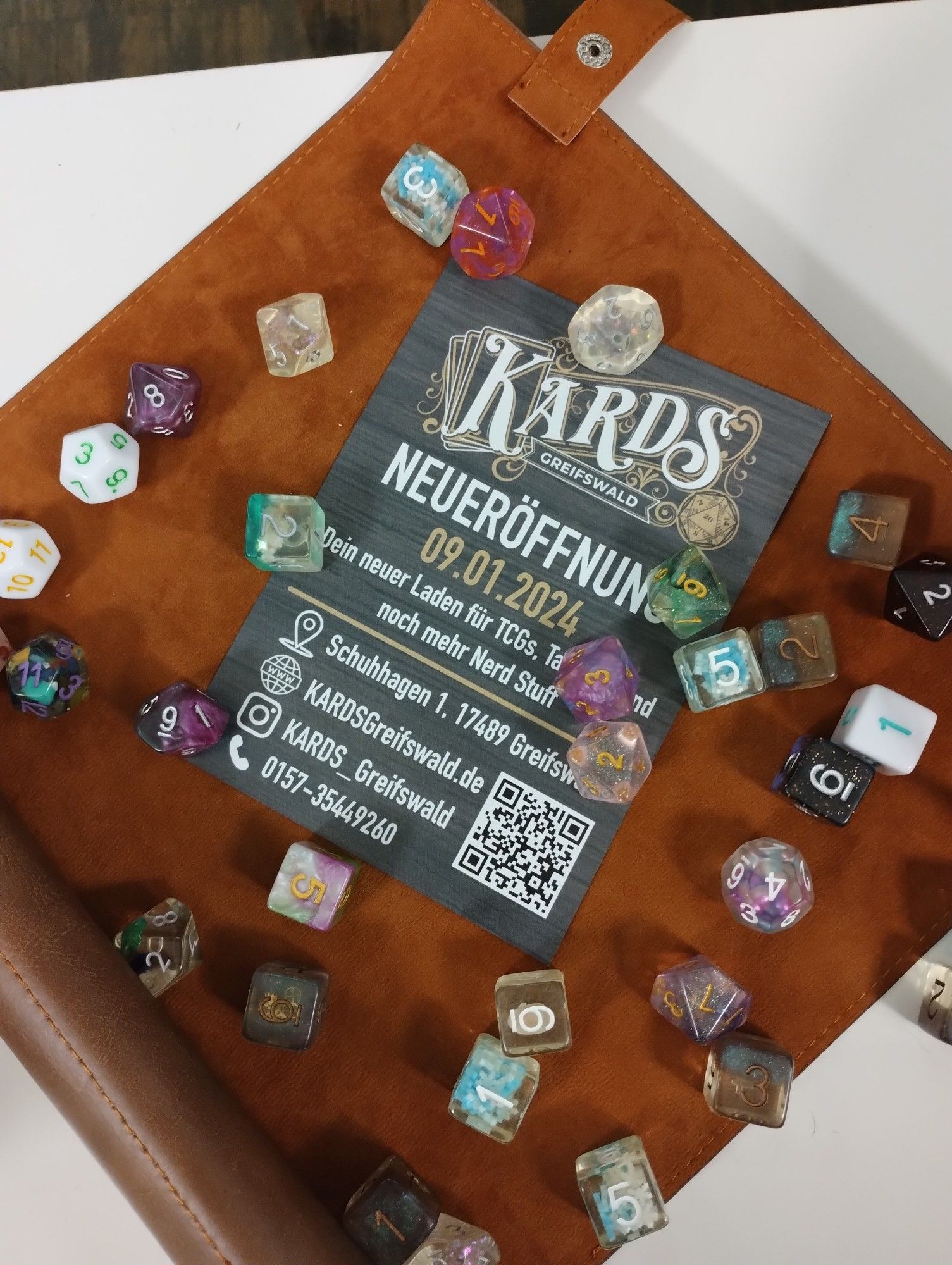 Ein Flyer mit Würfeln, die drum herum liegen. Auf dem Flyer steht "KARDS Greifswald - Neueröffnung 9.1.2024. dein neuer Laden für TCGs, Tabletop und noch mehr Nerd Stuff. Schuhhagen 1, 17489 Greifswald. Web: kardsgreifswald.de Instagram: KARDS_Greifswald Tel 015735448260"