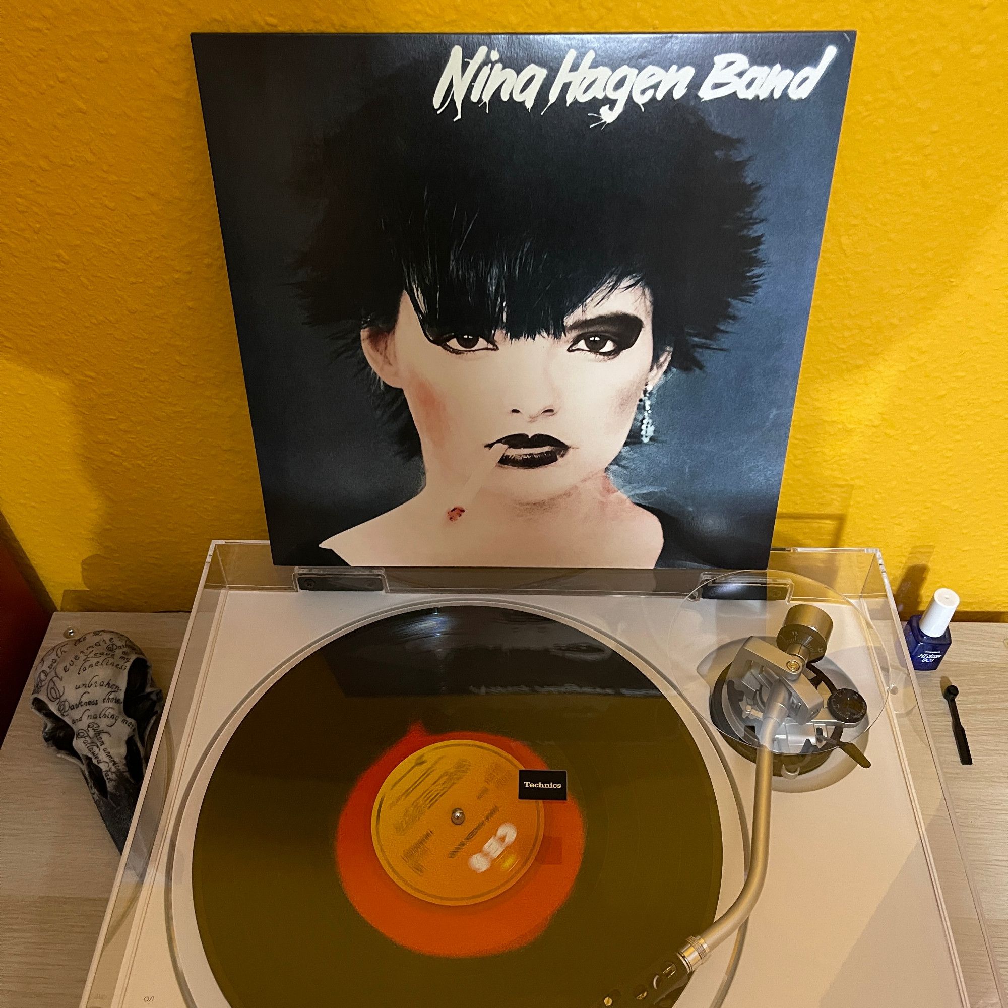 Blick von schräg oben auf den weißen Plattenspieler mit geschlossener Haube, auf der die Plattenhülle steht und gegen die gelbe Rückwand lehnt. Auf dem Cover ist ein Portrait von Nina Hagen mit Kippe im Mundwinkel, die Farben sind sehr kontrastreich gehalten. Das Vinyl auf dem Plattenteller ist schwarz mit einem transparent roten Fleck in der Mitte.