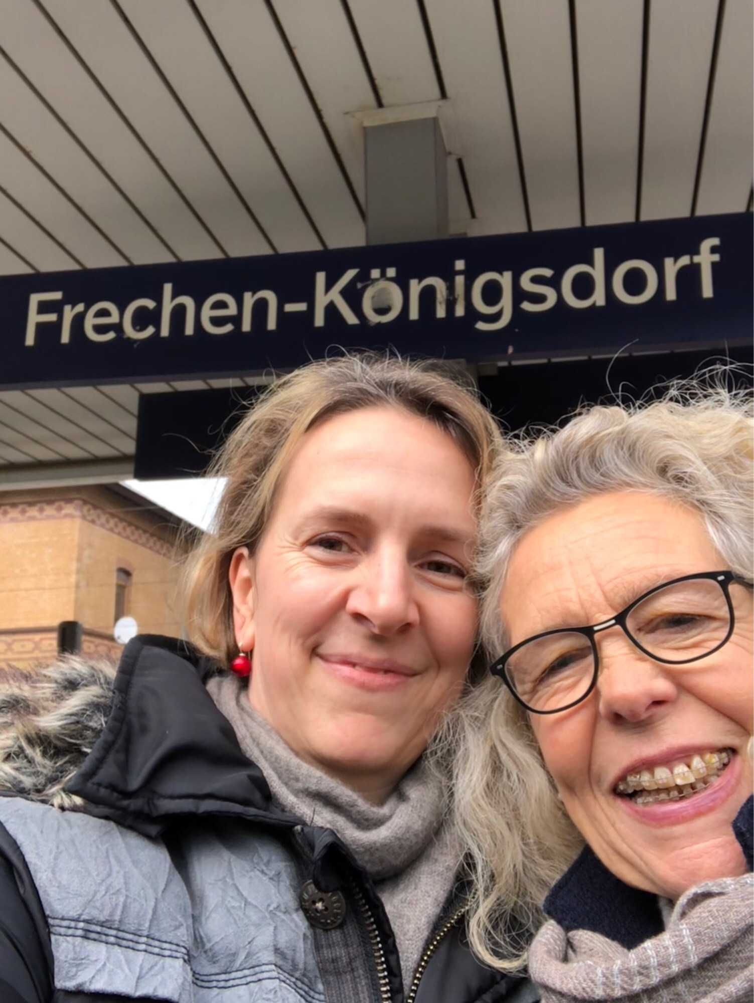 Die Autorin des Beitrags  (Dagmar Karrasch) und Antje Krüger machen ein gemeinsames Selfie vor einem Bahnhofsschild mit der Aufschrift Frechen-Königsdorf. Beide freuen sich aneinander.