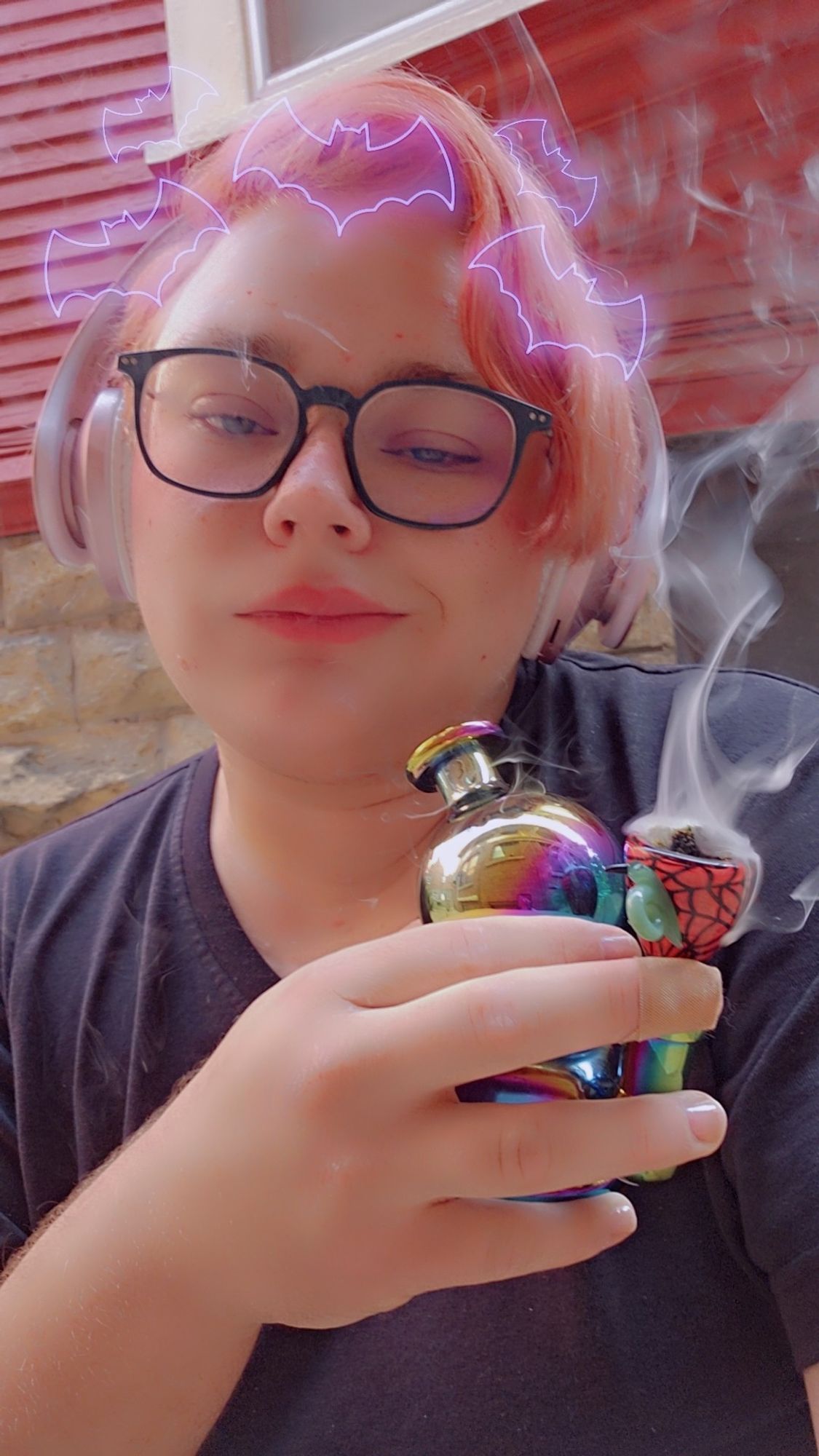 A Snapchat image of Arden with purple bat outlines around his head.Arden is wearing a pair of pink and white headphones and seated outside. Arden's eyes are half lidded, and it is holding a small iridescent bong that has smoke coming from the bowl. Arden is smirking at the camera while holding his lungs full of smoke.