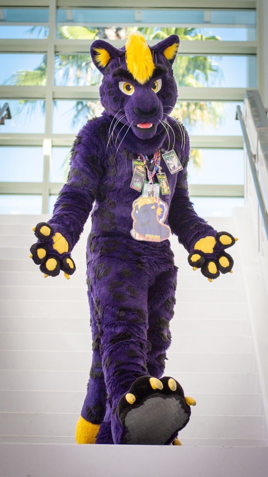 A purple and yellow panther fursuiter posing with his paws to the camera on a staircase