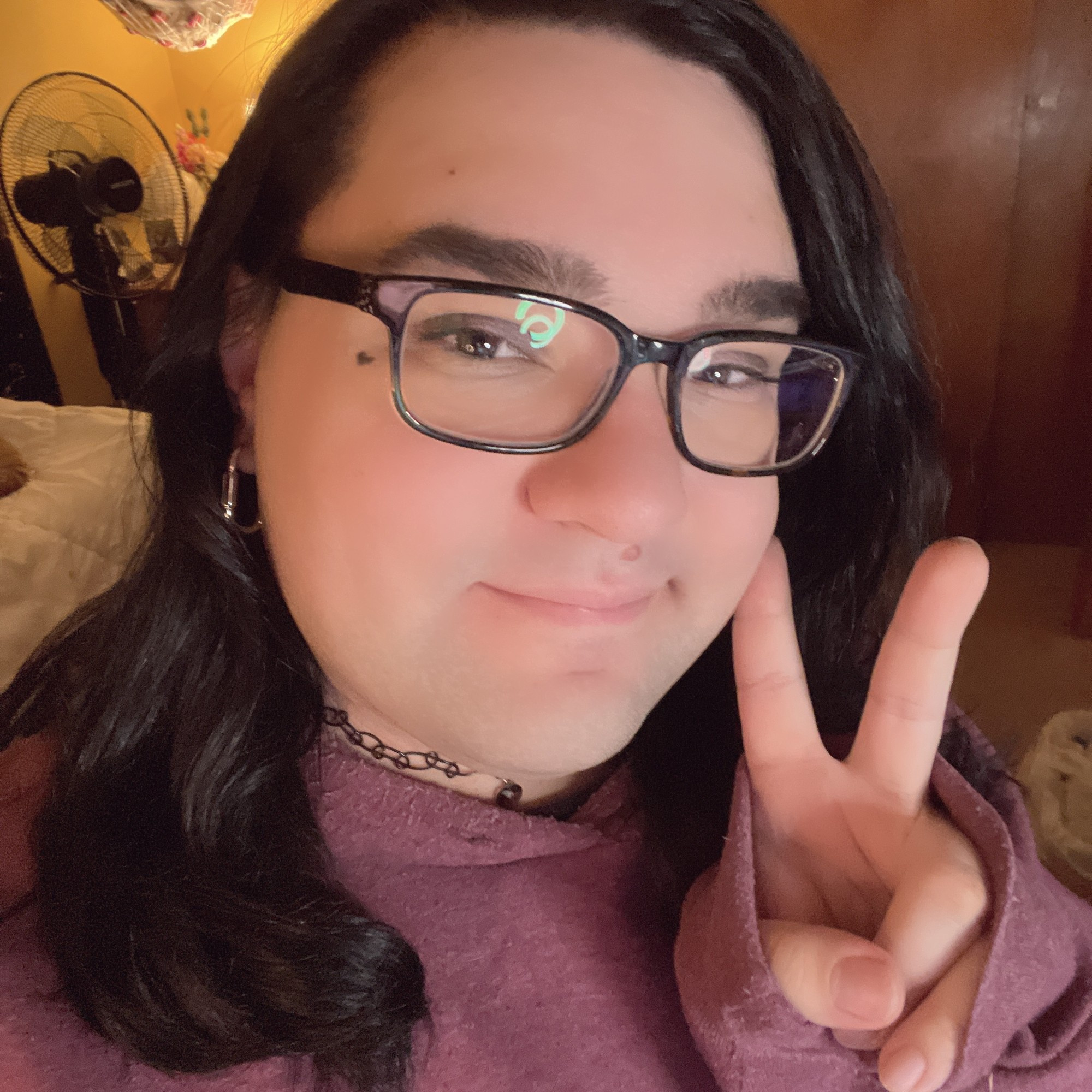 A selfie of me, Mei, in real life! Shows me, doing the peace sign, with medium-length black hair, square glasses with a small heart next to my left eye, wearing a unsaturated burgundy red hoodie with a black tattoo choker and a lock earring!