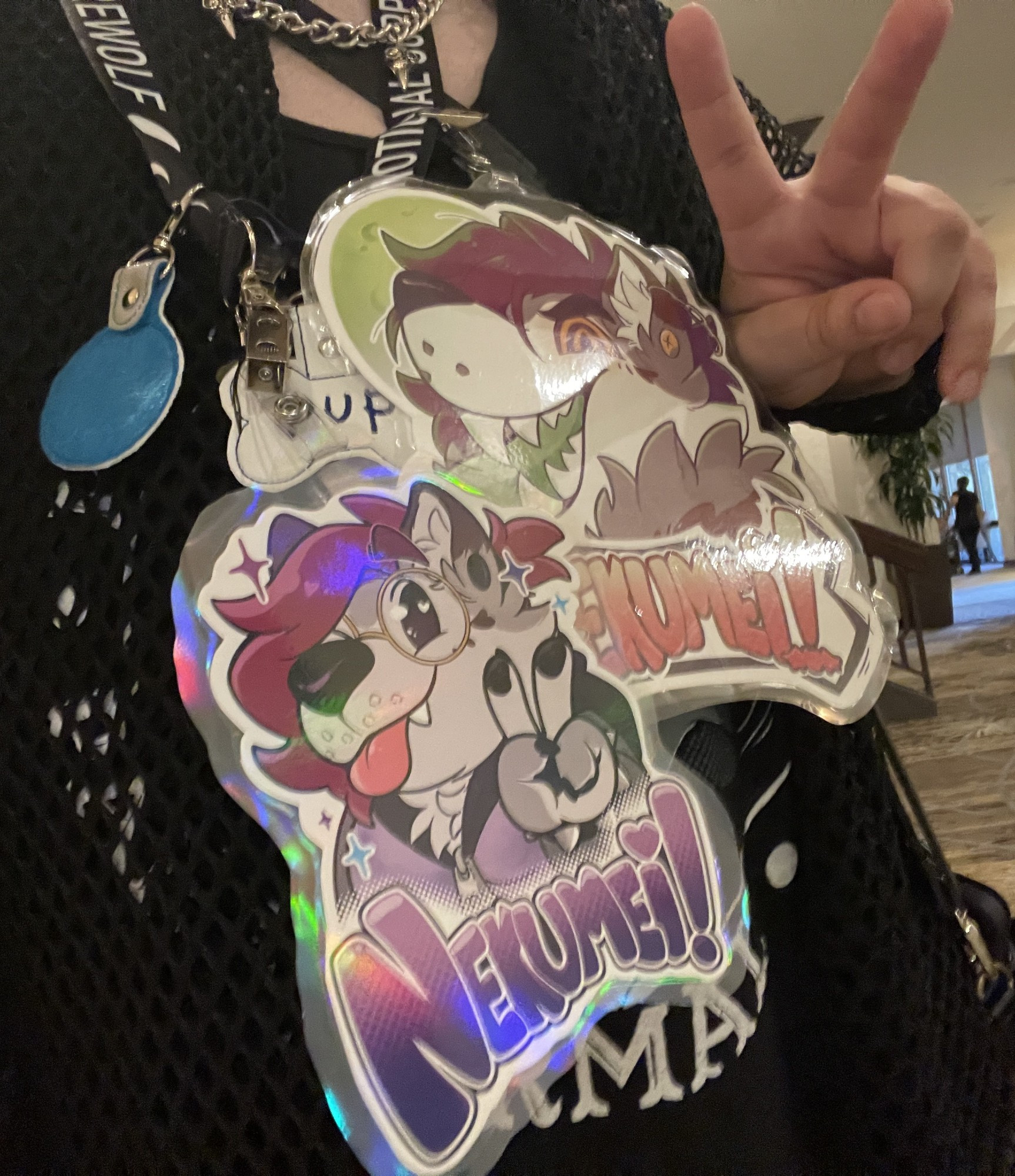 A picture of me wearing my various con badges I'll be showing at BLFC! The badge at the bottom reads "Nekumei!" in purple-gradient colors with my wolfdog fursona, Mei, doing the peace sign (like what I'm doing in the picture!), and the badge above it also reads "Nekumei" but in orange-yellow gradient text and features my were-wolfdog fursona Mei looking at the viewer and grinning menacingly