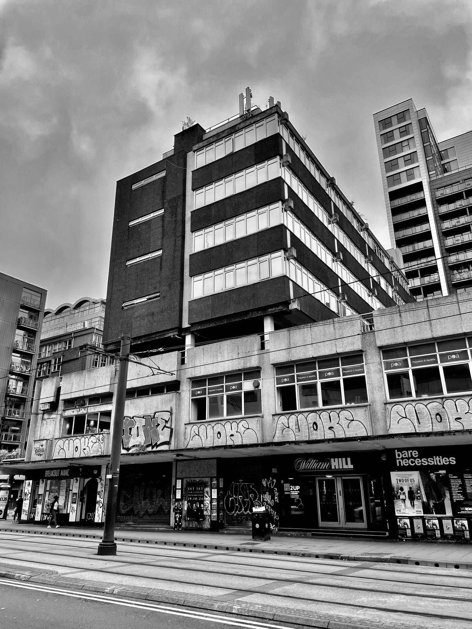 A black and white photo of a squat, graffiti-covered concrete block of a building. In the foreground are tram tracks and a pole for overhead wires.