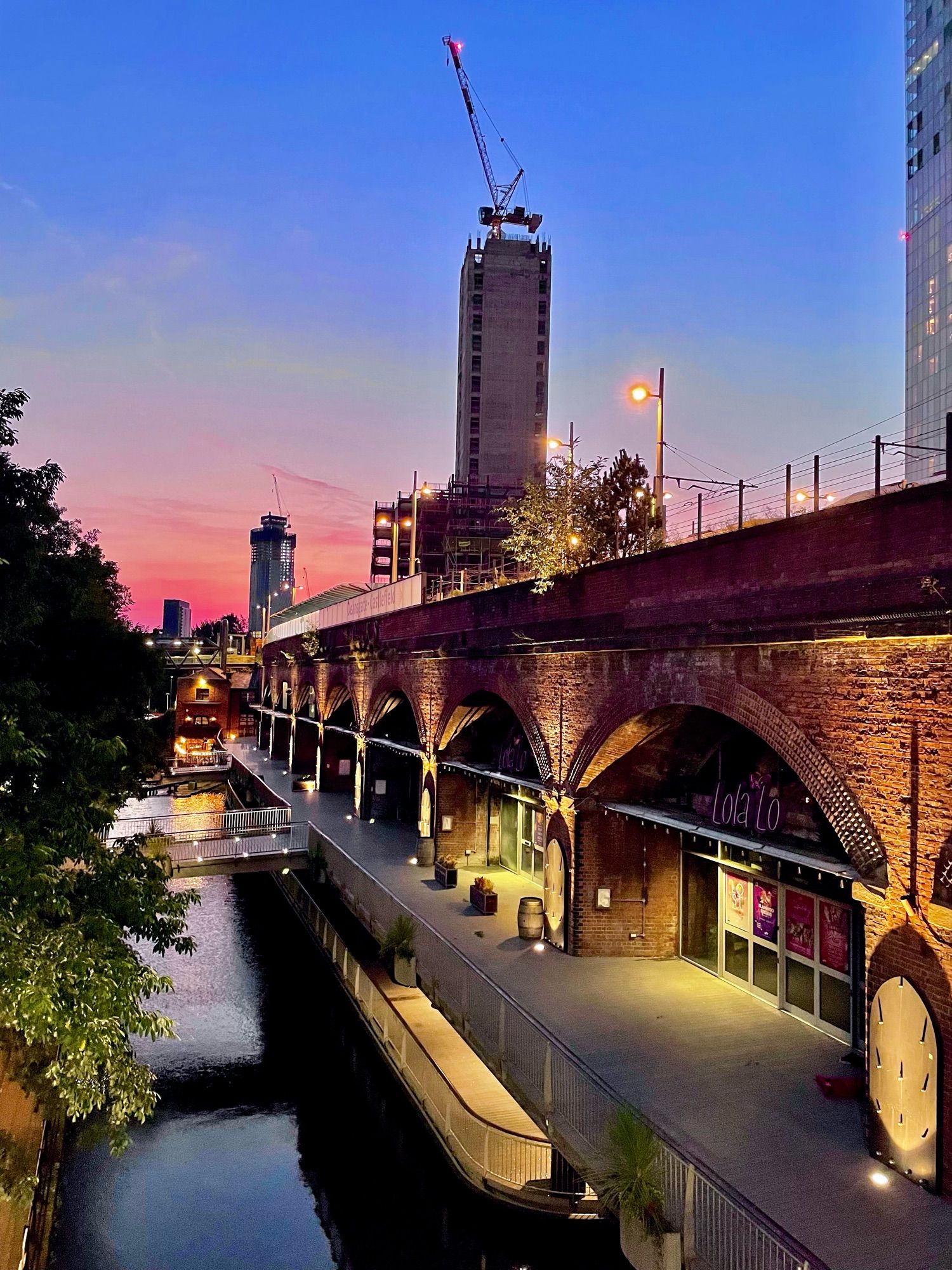 An elevated tram station above canal side vaults, built with red brick. In the background are a couple of high rise buildings and a crane; the sky is giving big bisexual lighting energy.