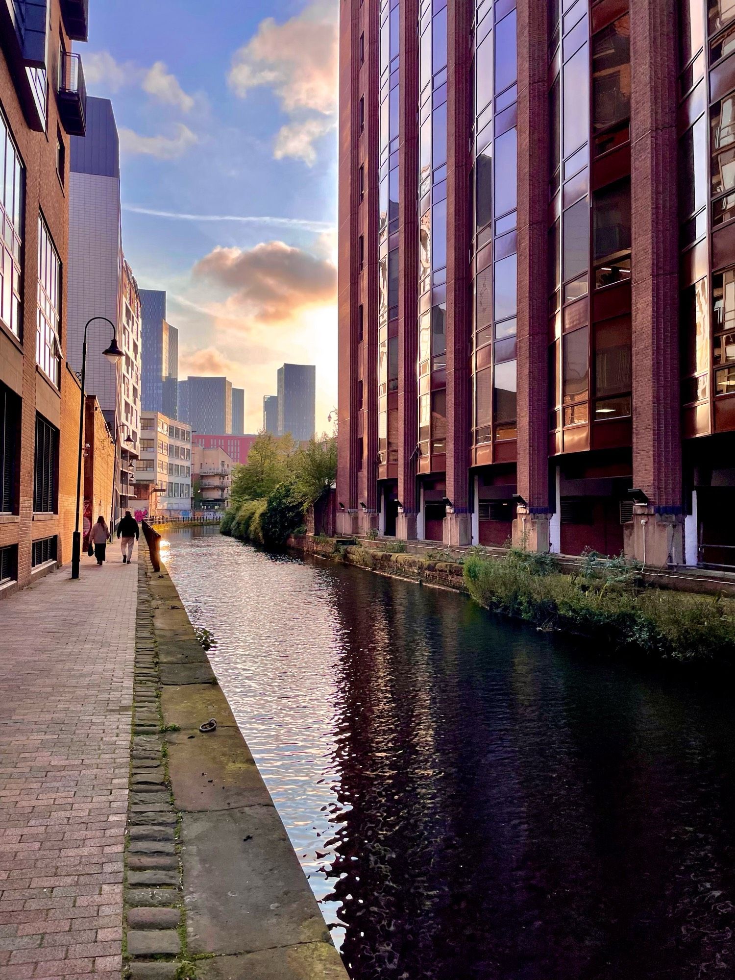 By the side of a canal, on both sides tall buildings in shades of red as the canal turns a corner at the photo horizon. The sky is cerulean blue with a couple of big fluffy clouds.