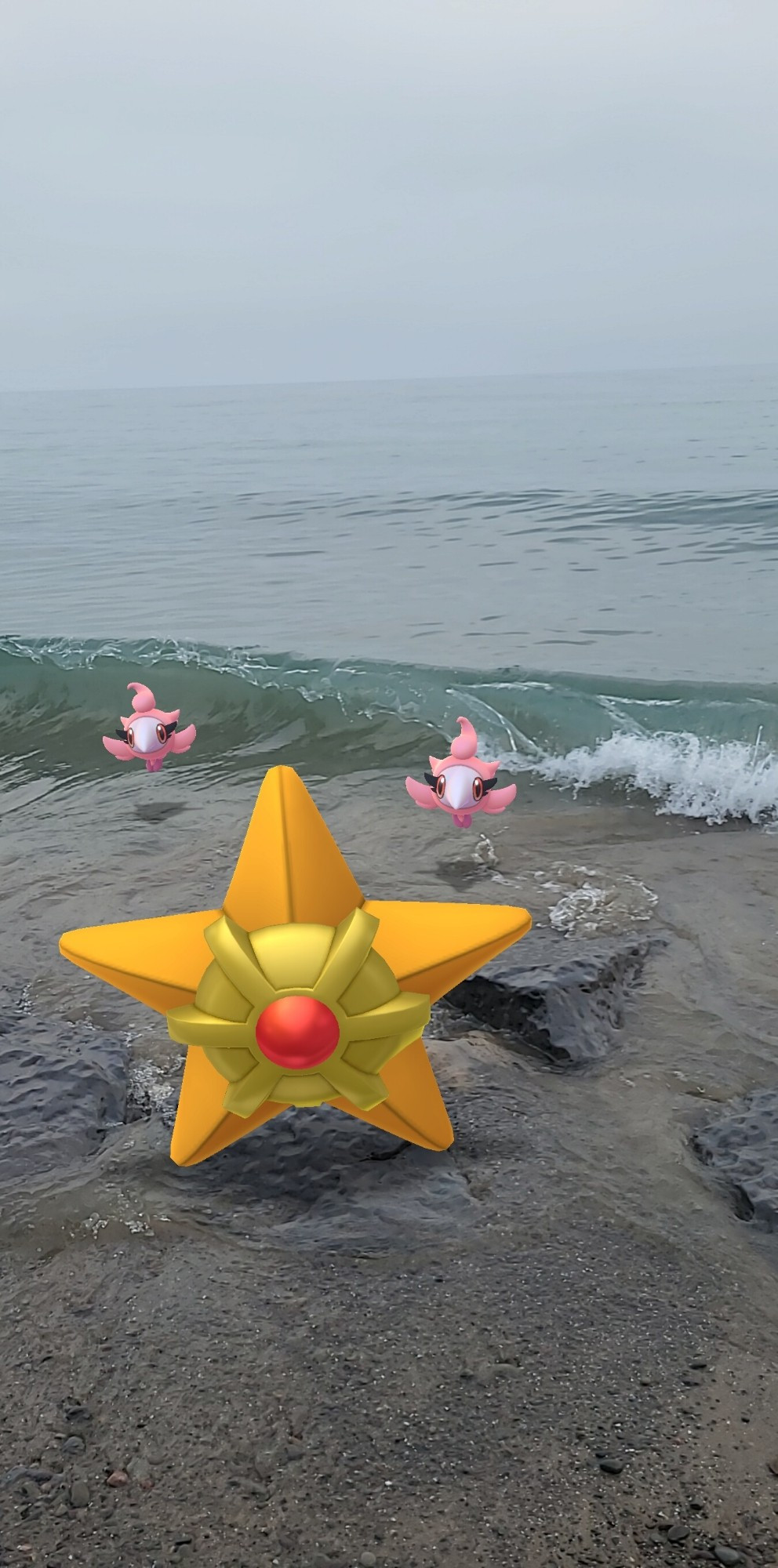 Staryu and two of the pink flying Pokemon, Spritzee. The pink Pokemon are standing right in front of a crashing wave, while Staryu stands on rocks submerged in the water. The water is a tealish blue while the sky is a grayish blue.