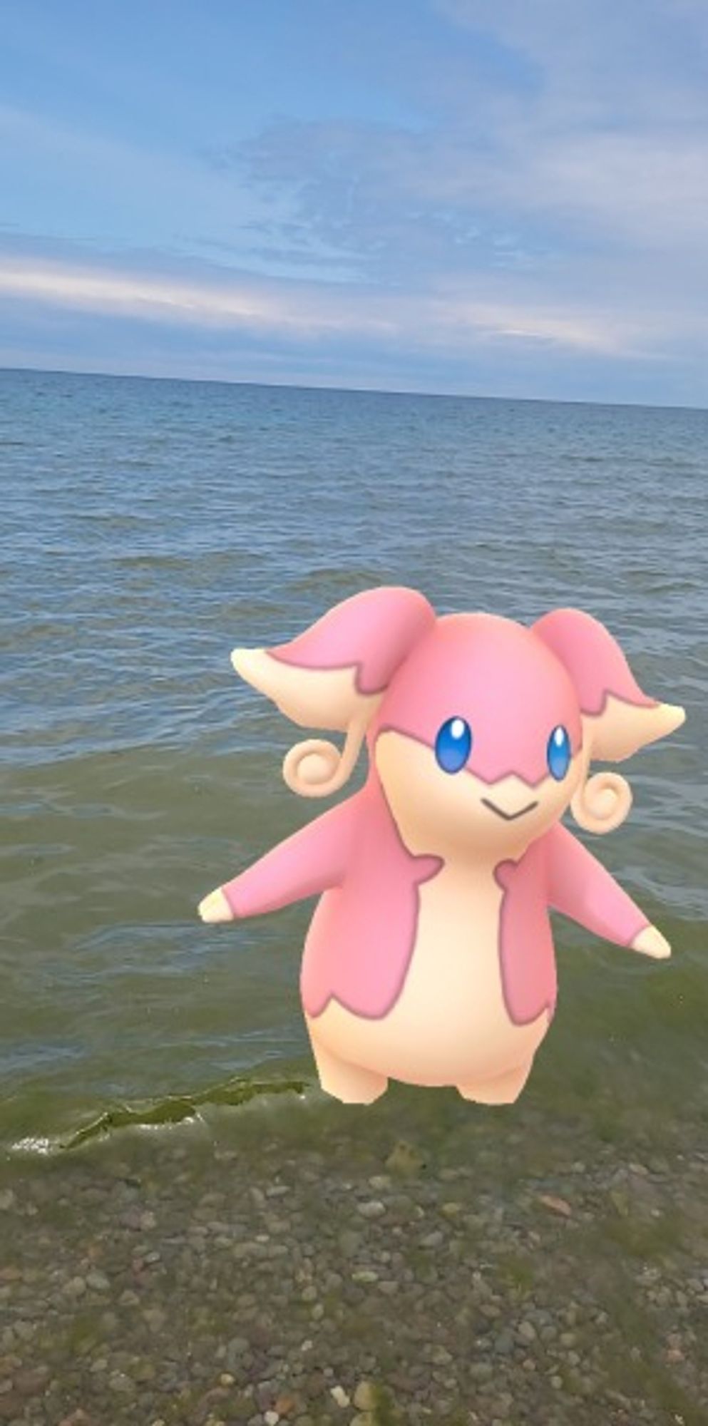 The Pokemon Audino wading on the shoreline, feet slightly submerged into the water. Silky white clouds make up the sky.