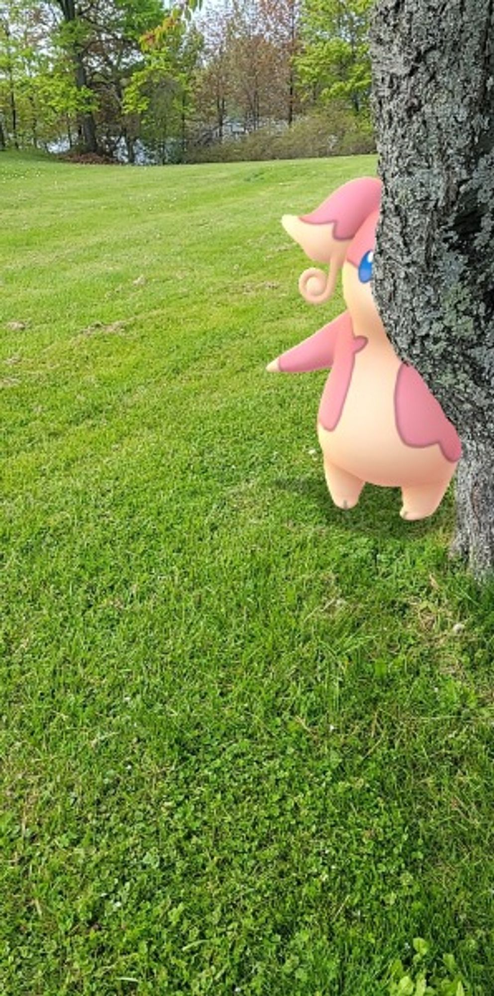 The Pokemon Audino hiding behind a tree.  Green grass makes up most of the foreground with a small assortment of green, brown and gray trees in the background with a blue lake barely visible behind it.