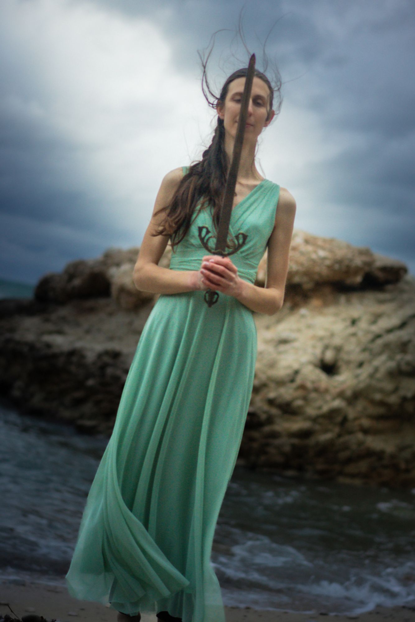 Photo of a woman on the coast, in a green dress, holding a sword 