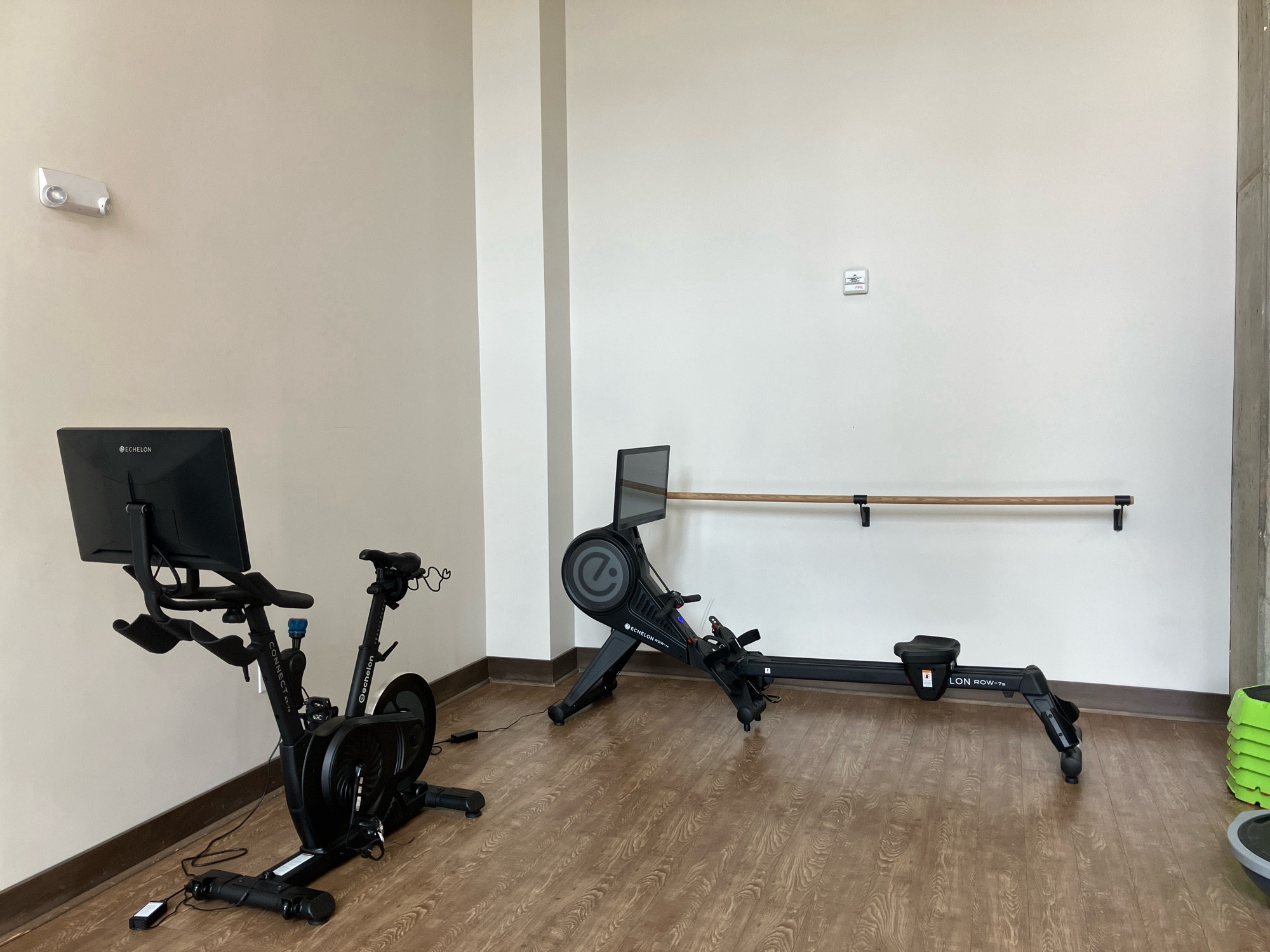 Photo of a gym shows spinny cycle along one wall with room at each end and no barre on the wall, another wall at 90 degrees has a mounted barre that is inaccessible because the rowing machine placed along it is nearly as long as the barre is. The spinny cycle is much shorter than the rowing machine.