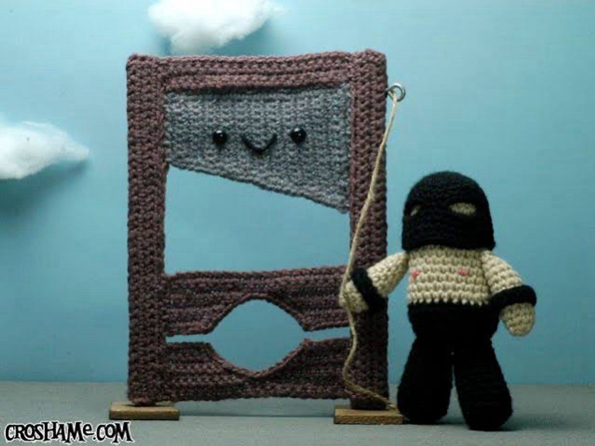 A crocheted smiling guillotine being prepared for its very useful function by a tiny crocheted operator