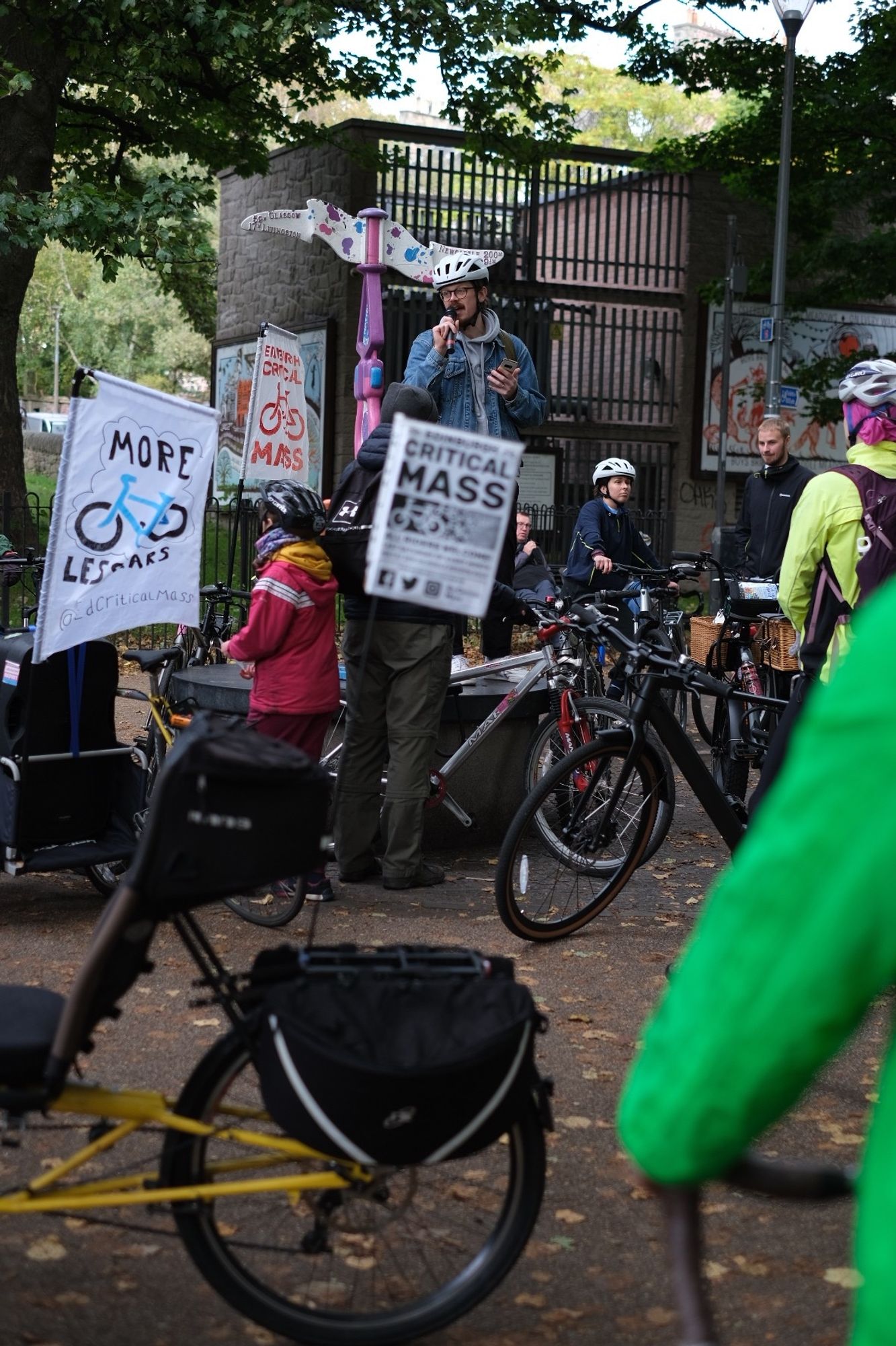 A large group of cycling enthusiasts stand together in front of a man in a denim jacket and bicycle helmet with a microphone.  There's a official cycle path marker behind him.