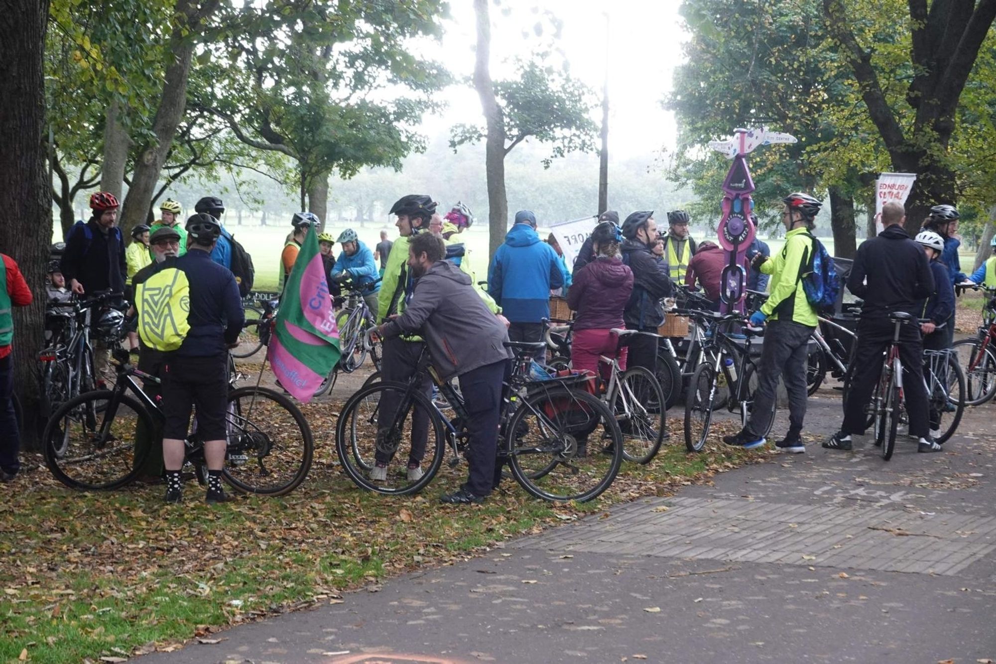 Lots of cyclists stand around in a park - many have flags with slogans promoting active travel