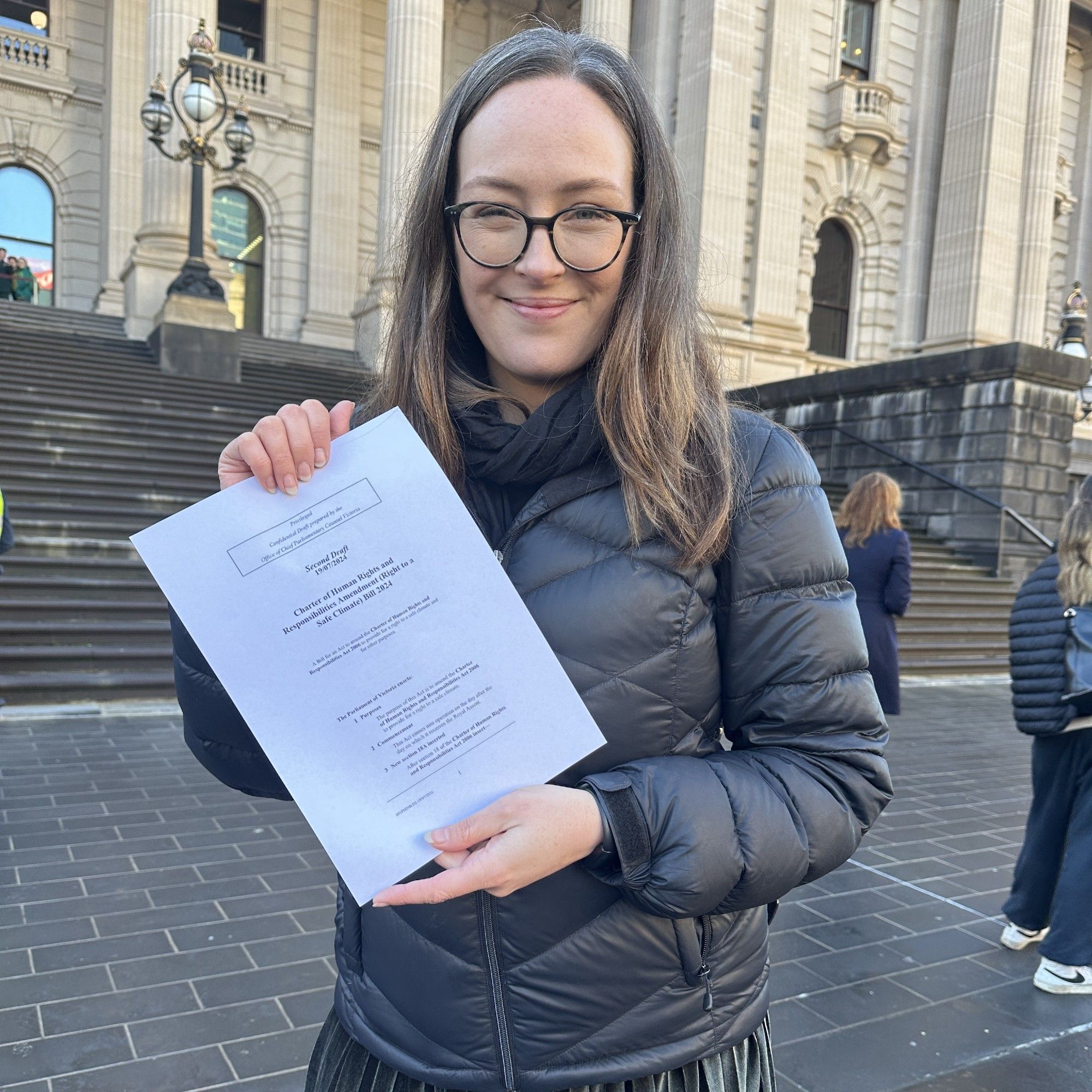 Katherine Copsey MP stands in front of the Victorian parliament holding the bill to add a right to a safe climate into Victoria's Charter of Human Rights