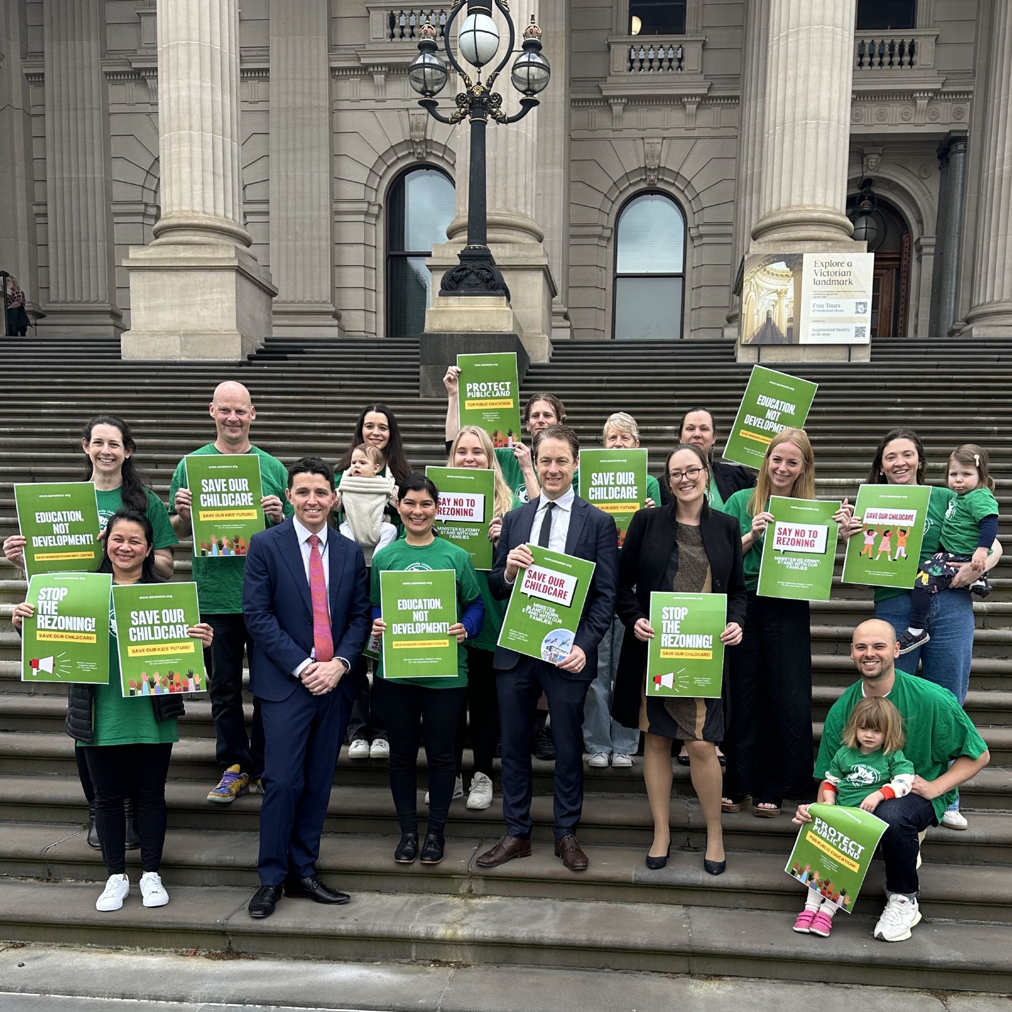 A group of people in green t-shirts hold green signs with slogans like "Education, not development!", "Save our childcare", "Stop the rezoning" and "Protect public land". Amongst this group are three MPs, Ryan Batchelor, Sam Hibbins and Katherine Copsey. All are standing on the steps of the Victorian Parliament.