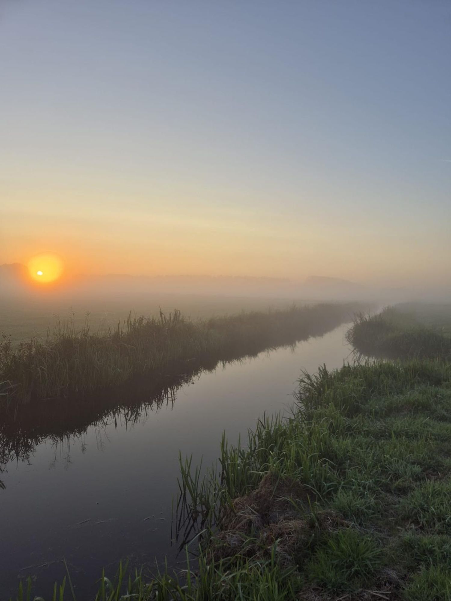 Opkomende oranje zo'n.met mist over ene weiland met een sloot en groen gras