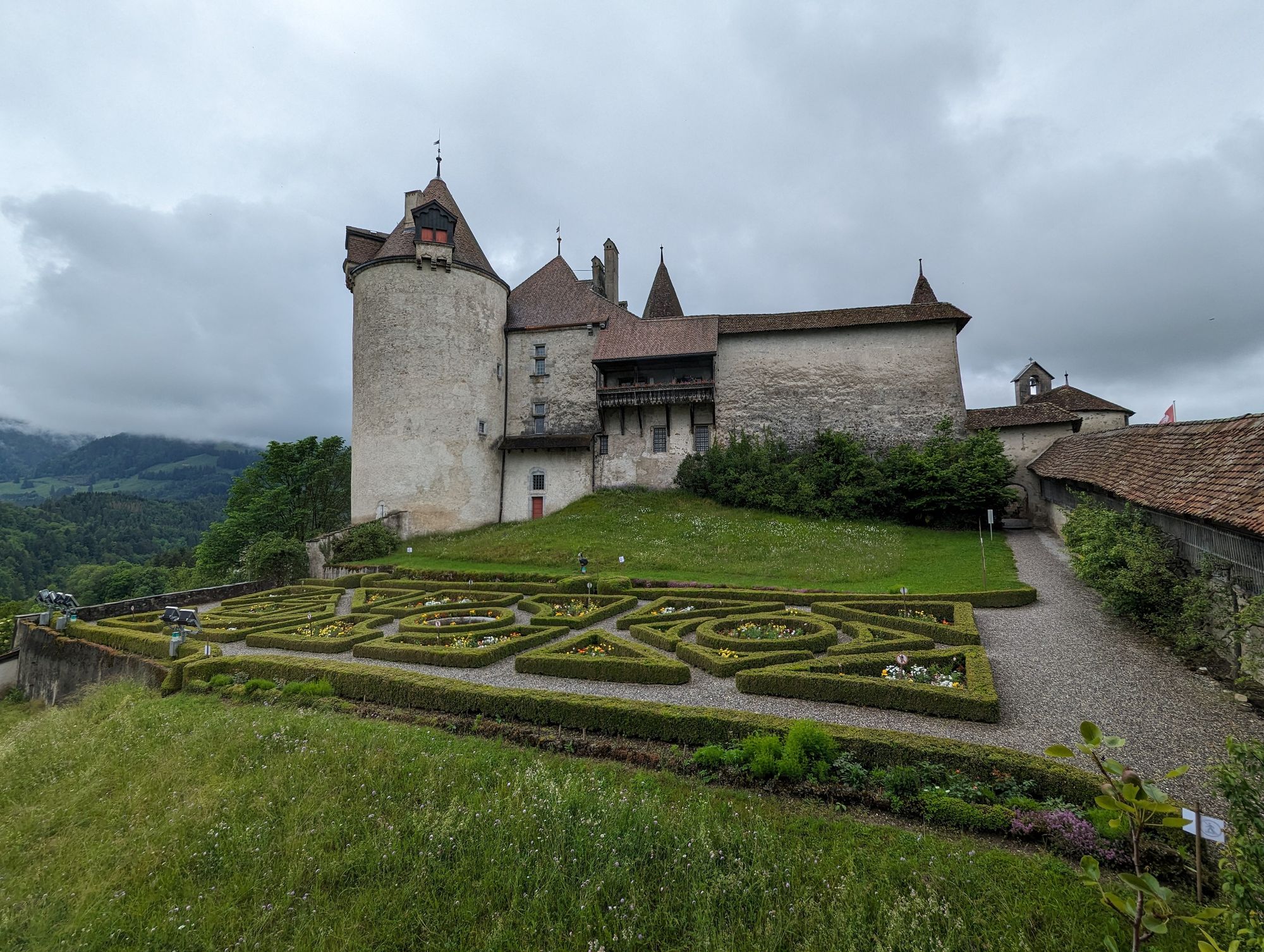 Gruyère castle. The front of the photo is filled by a manicured French flower garden, with small box hedges. In the back, an old castle looms. To the side, a view is opening into the green valley below. There is a heavy cloud cover overhanging.