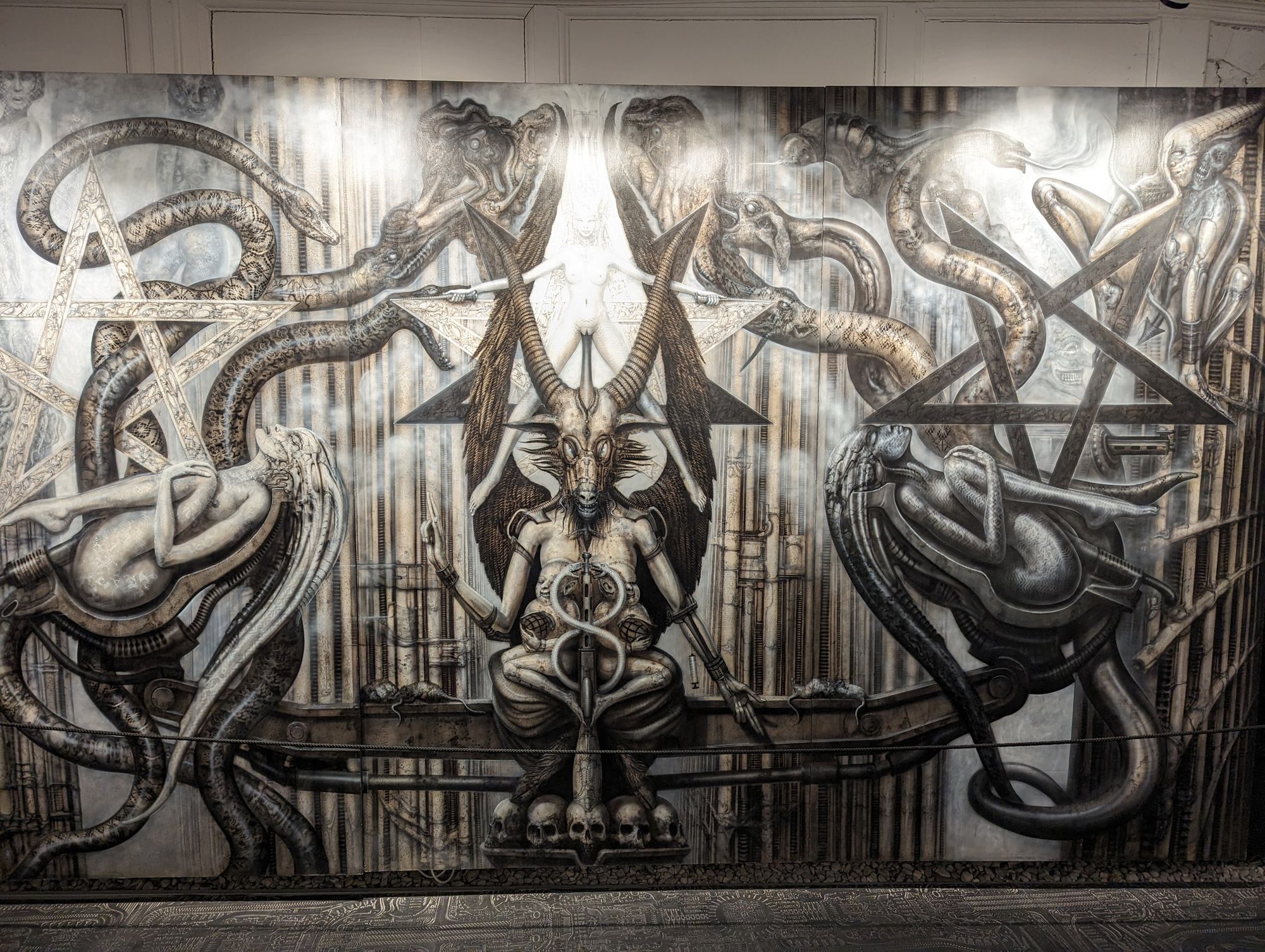 A black and white HR Giger painting, covering a whole wall. In the center, Lucifer sits, a naked woman behind him. He's flanked by pentagrams and snakes and more naked women. On closer view, they all sit on machines that seem to be copulating with them. There's skulls and dead rats.