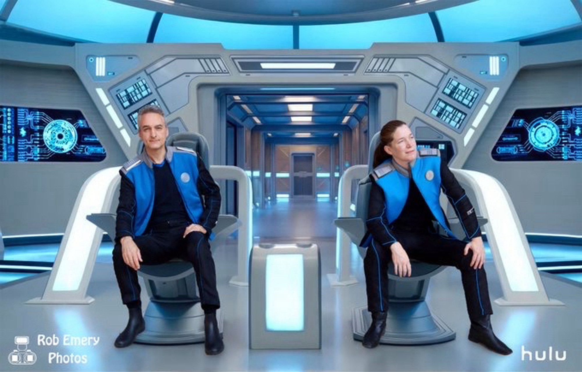 Two Orville cosplayers, both white and in command blue with open jackets, sitting on the bridge.