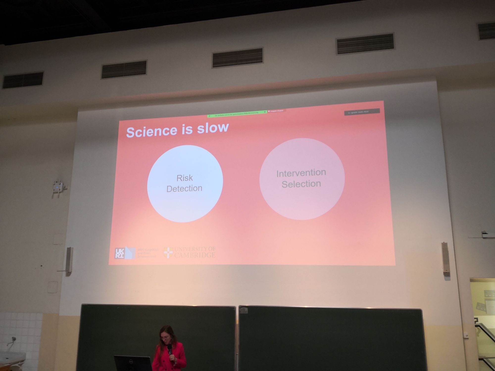 Photo of Amy Orben in front of a presentation. The slide says: Science is slow. Risk detection. Intervention selection.