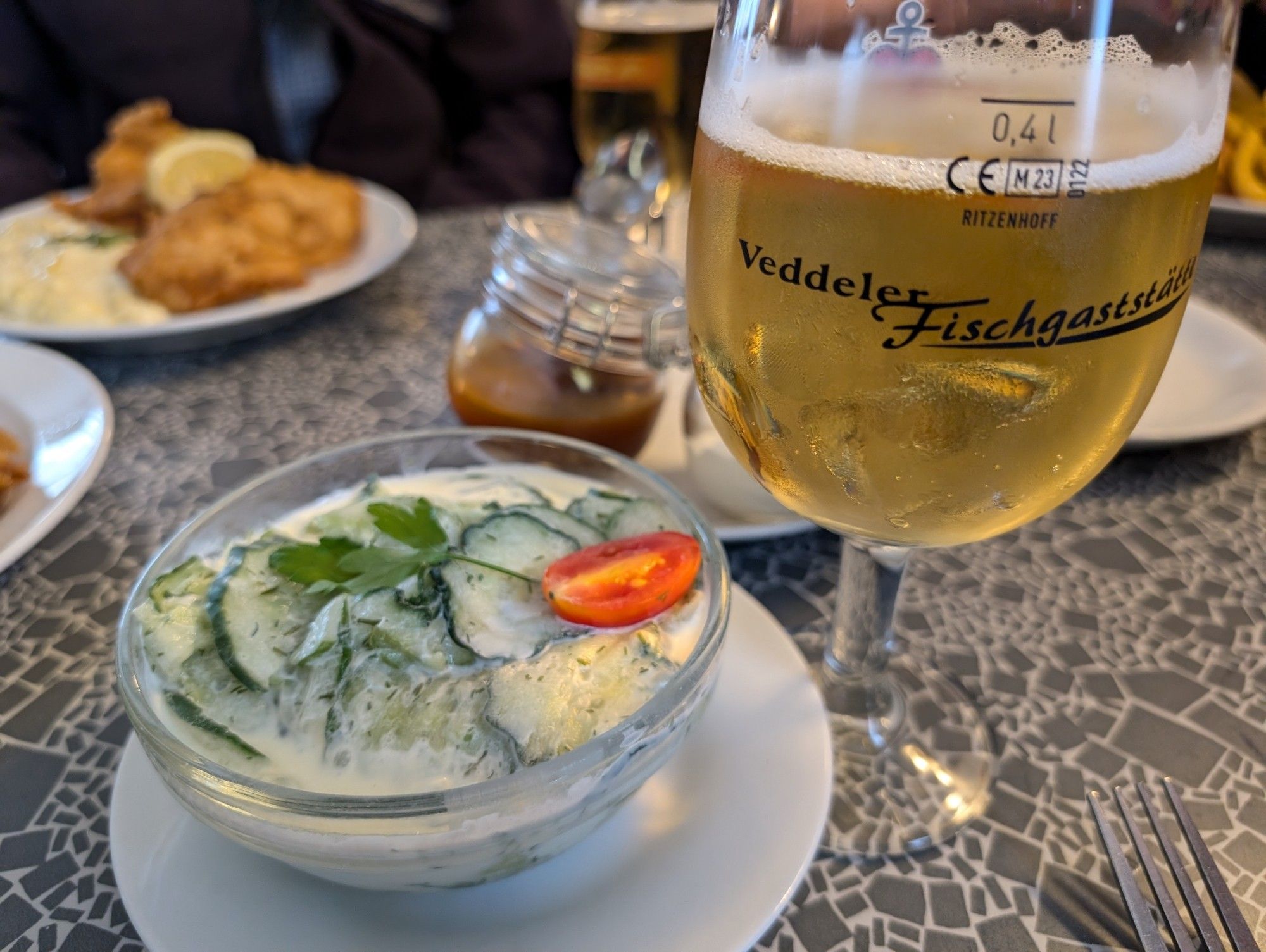 Ein mit Alsterwasser gefülltes Bierglas mit der Aufschrift "Veddeler Fischgaststätte" steht neben einem Gurkensalat, im Hintergrund sind ein Glas mit Ketchup und ein Fischgericht zu sehen