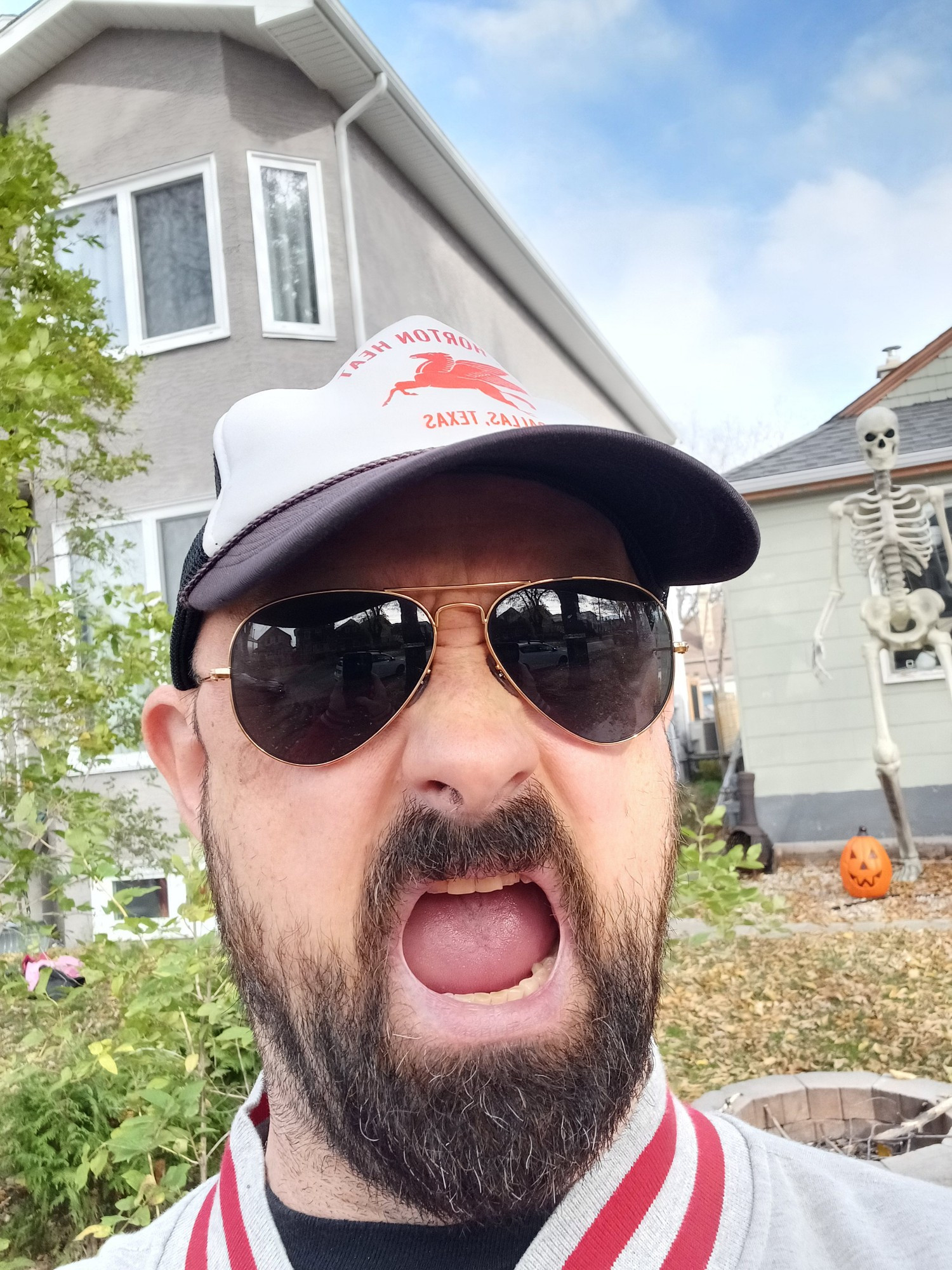 A selfie with me in the foreground (a white male with a brown beard wearing sunglasses and a Reverend Horton Heat trucker cap, my mouth open wide in feigned alarm) and a giant Home Depot skeleton in the background. 