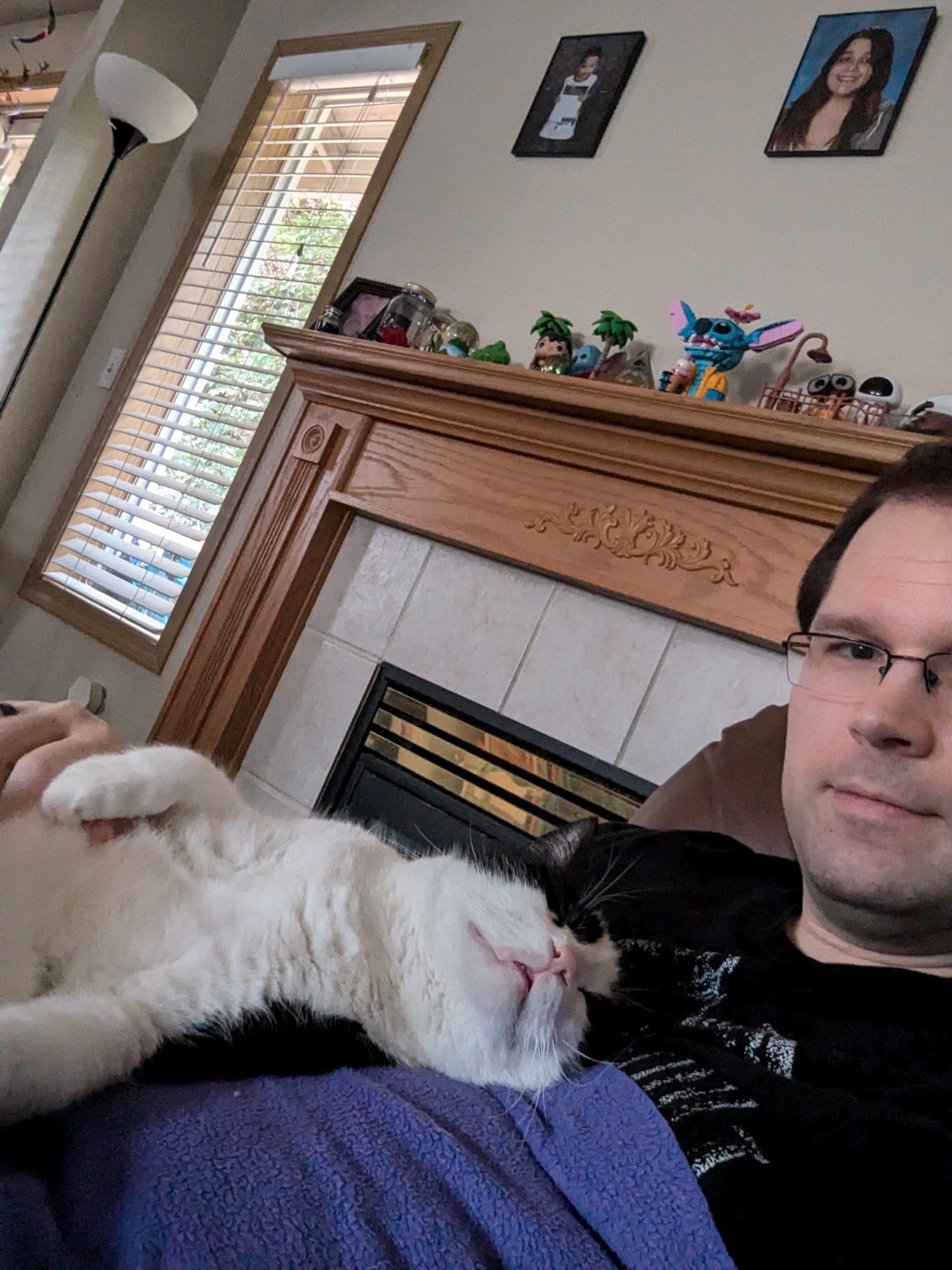 A black and white cat laying on his human with his white belly fur facing upward. The human is covered by a purple blanket. The cat appears to be napping with a look of comfy contentment on his face.