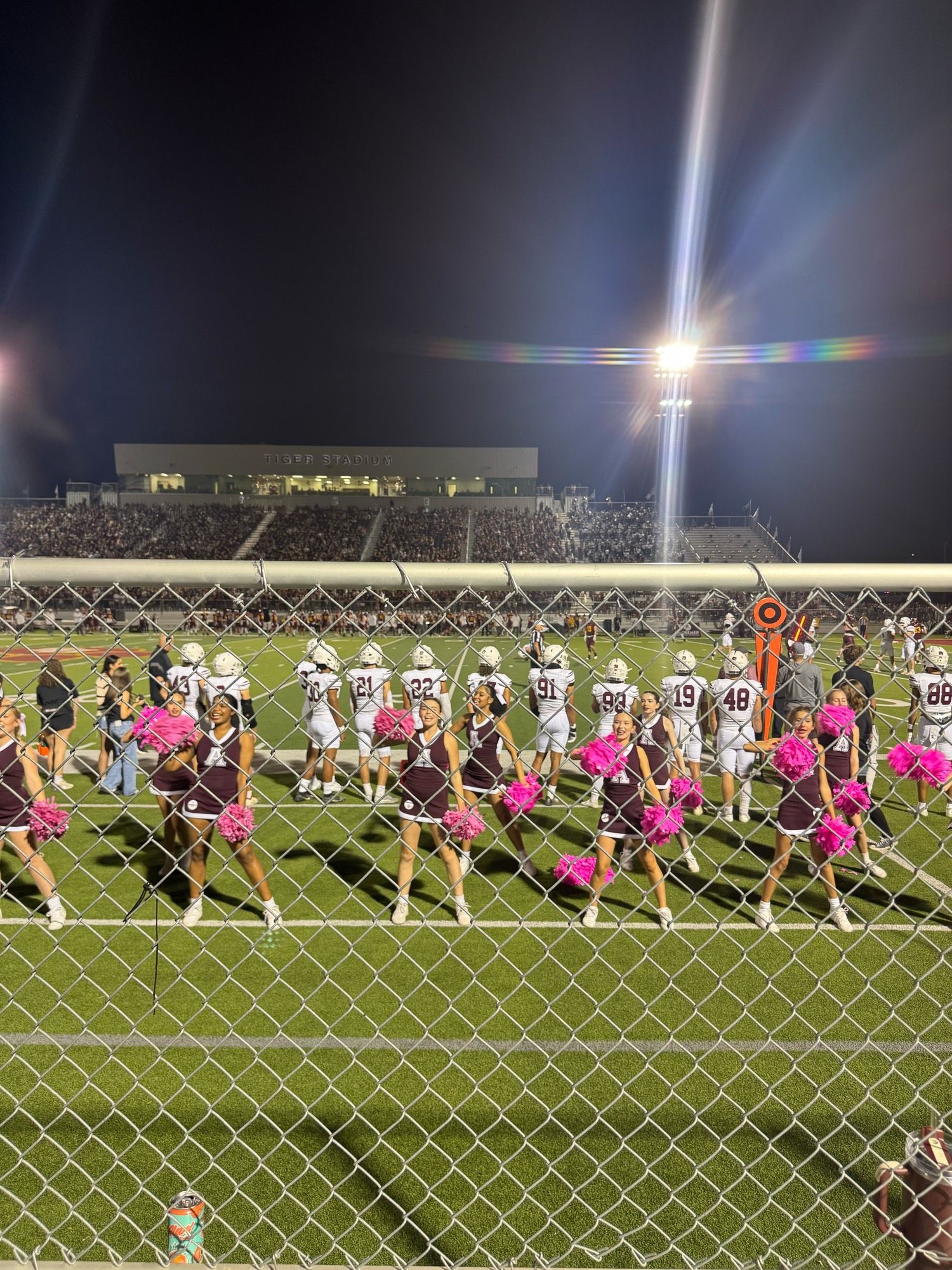 Through a chain link fence, cheerleaders and football players and a full stadium and a Friday night light flaring. An Arizona tea on a ledge in the foreground.