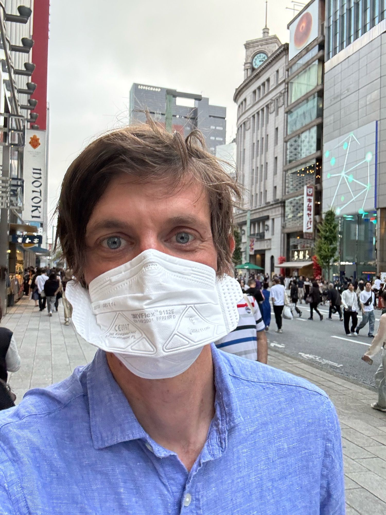 Michael Osborne takes a selfie on a busy Tokyo street. He wears a white 3M VFlex mask and a light blue button-down shirt. Behind him, crowds of people walk along a shopping district with tall buildings, including one with a clock tower.