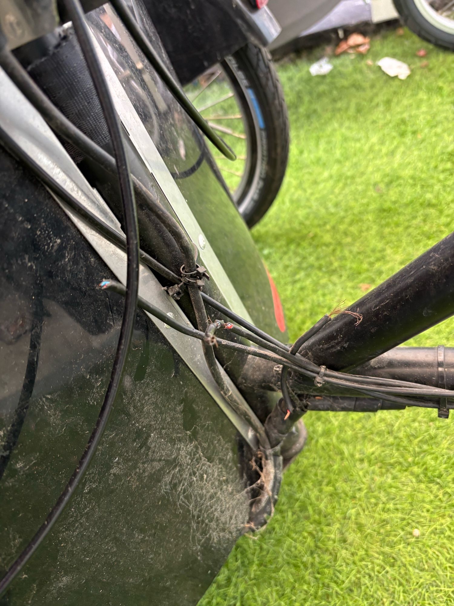 The image shows a close-up view of a bicycle frame, focusing on a section where several cables and wires are visibly damaged. The cables appear to have been chewed through, with frayed ends and exposed wires visible. This damage is consistent with the earlier mention of a dog chewing on power cables of a cargo bike.

The bicycle frame is black, and parts of a wheel and other bike components can be seen in the background. The bike is positioned on what looks like artificial grass or a lawn, suggesting it's outdoors.