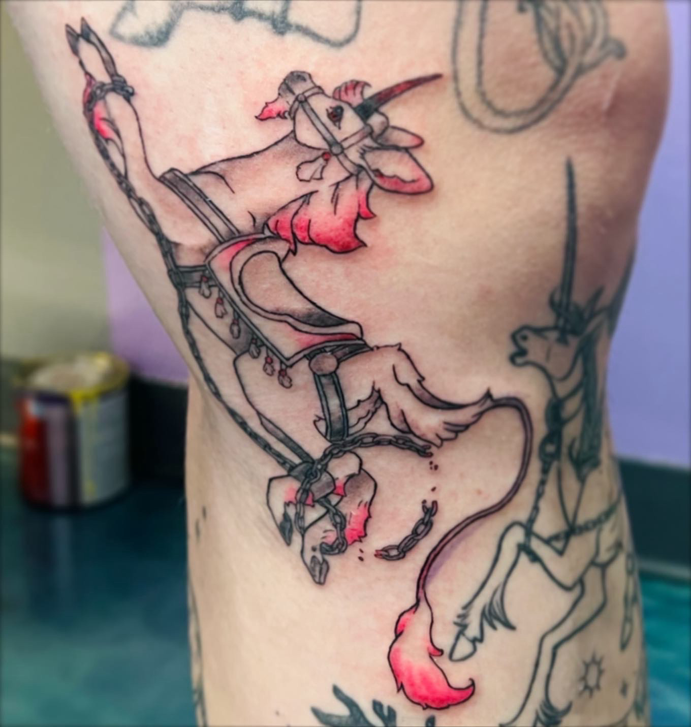 tattoo of a unicorn on clients leg- it’s wearing carousel style barding and is done in black and grey with hot pink accents on the mane and tail.