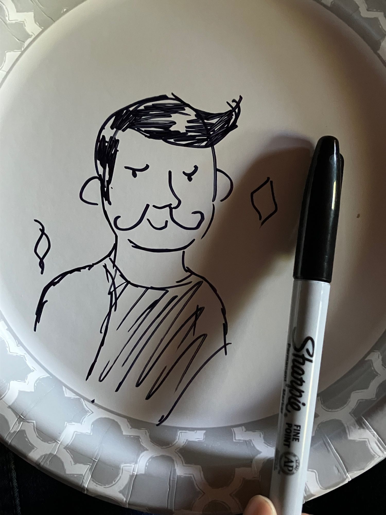 Marker drawing of Dorian simplified like a Peanuts character surrounded by sparkles on a paper plate. Sharpie is held next to the drawing