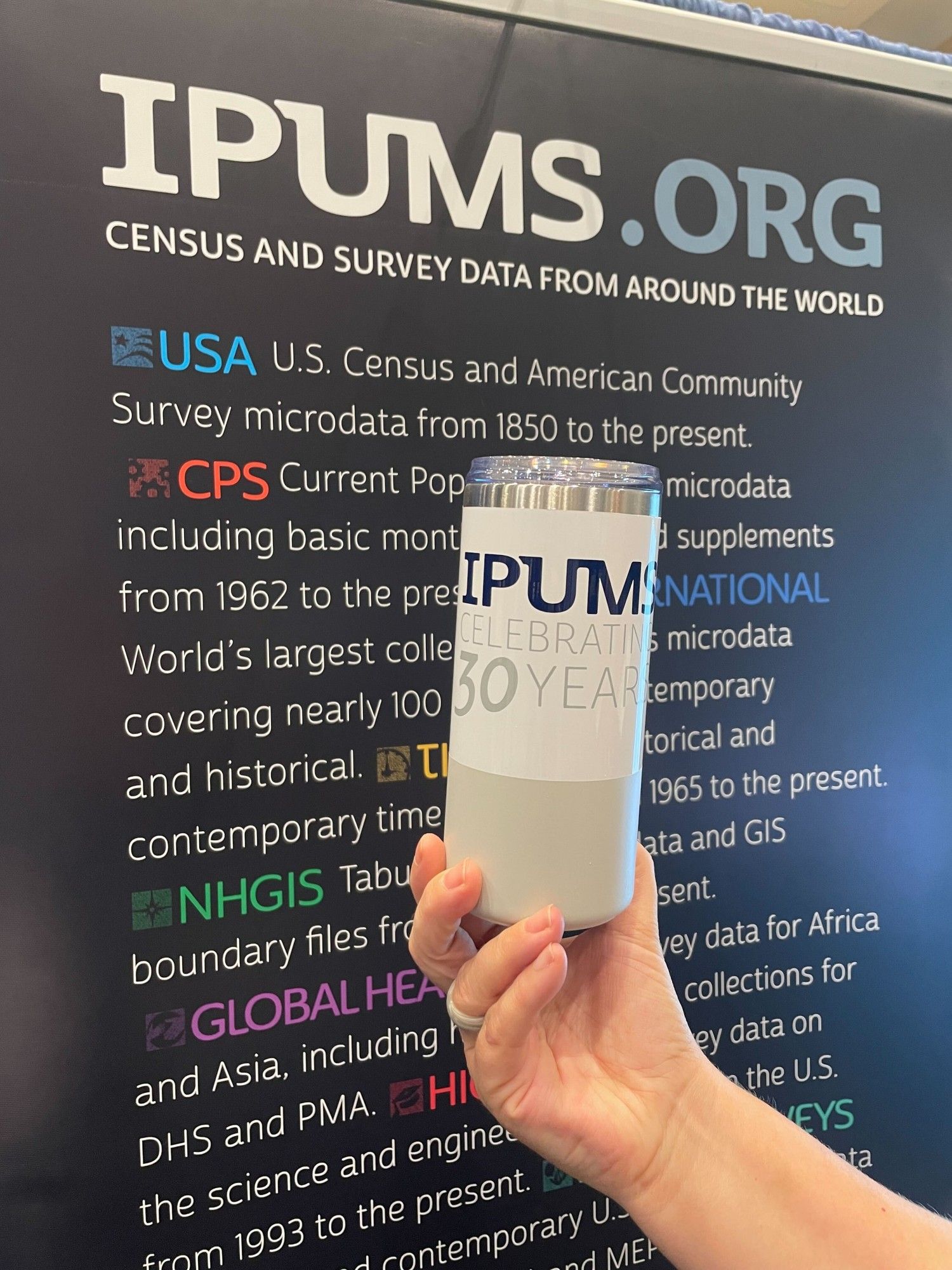 Picture of a person holding up a white and grey tumbler with the IPUMS logo on it in front of a banner at the IPUMS booth at PAA 2024. Come and claim yours if your #poweredbyIPUMS poster won an award!