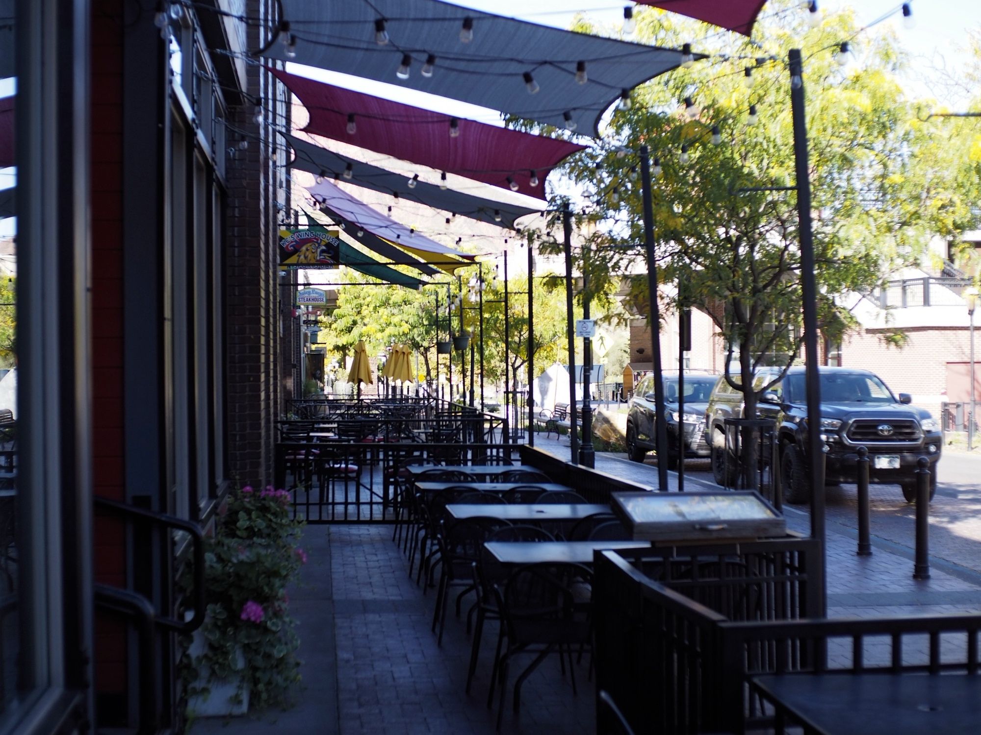 A series of dining patios, mostly hidden in shadow, with tress just beyond the darkness, bathed in sunlight.