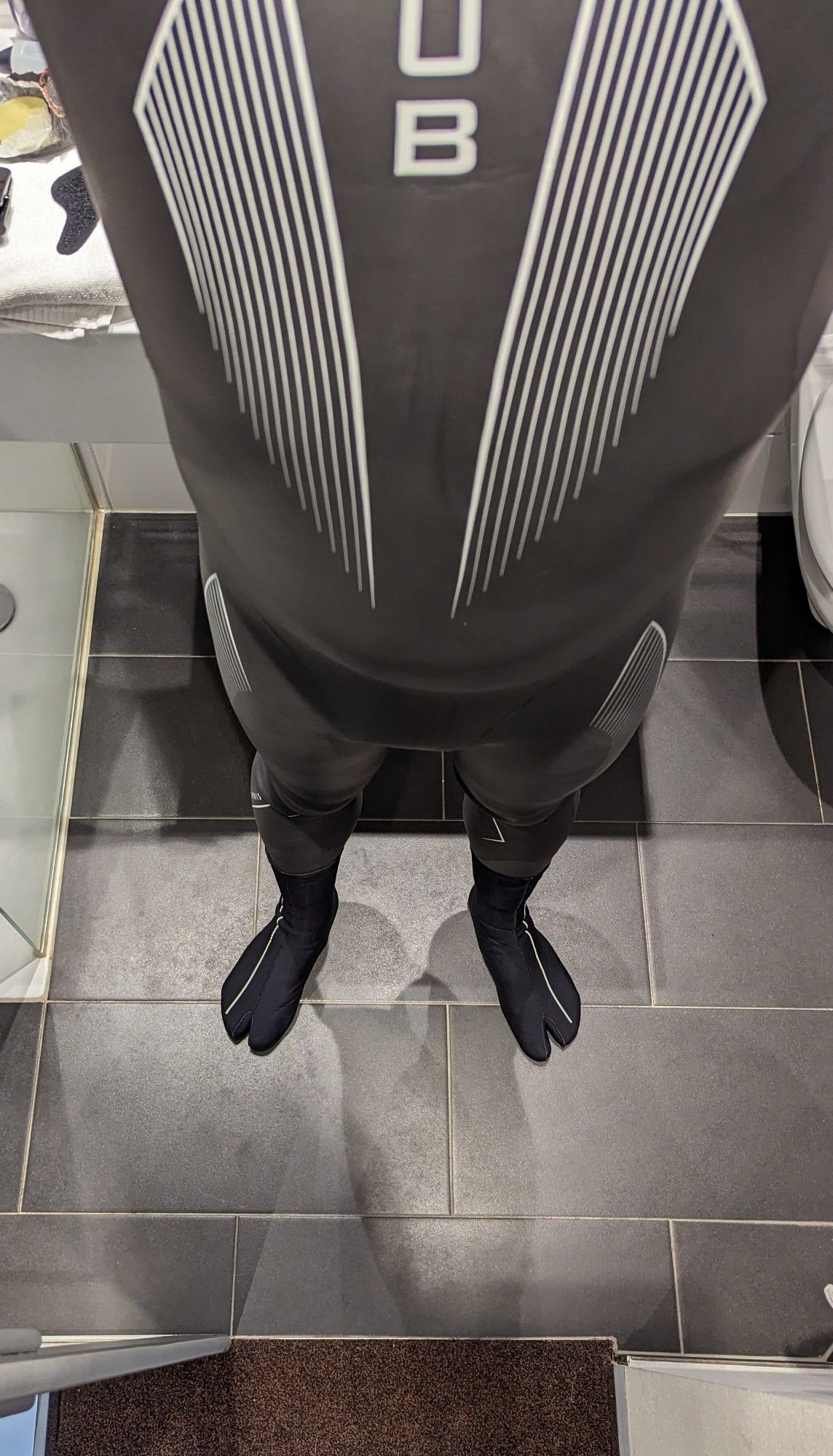 The image shows a person wearing a black wetsuit with white vertical stripes on the chest and legs, standing in a tiled bathroom. They are also wearing neoprene diving socks with a split toe design, likely for cold water activities such as surfing or diving. The wetsuit brand's logo, "HUUB," is visible on the chest area. The image is taken from the person's point of view, looking down at their body.