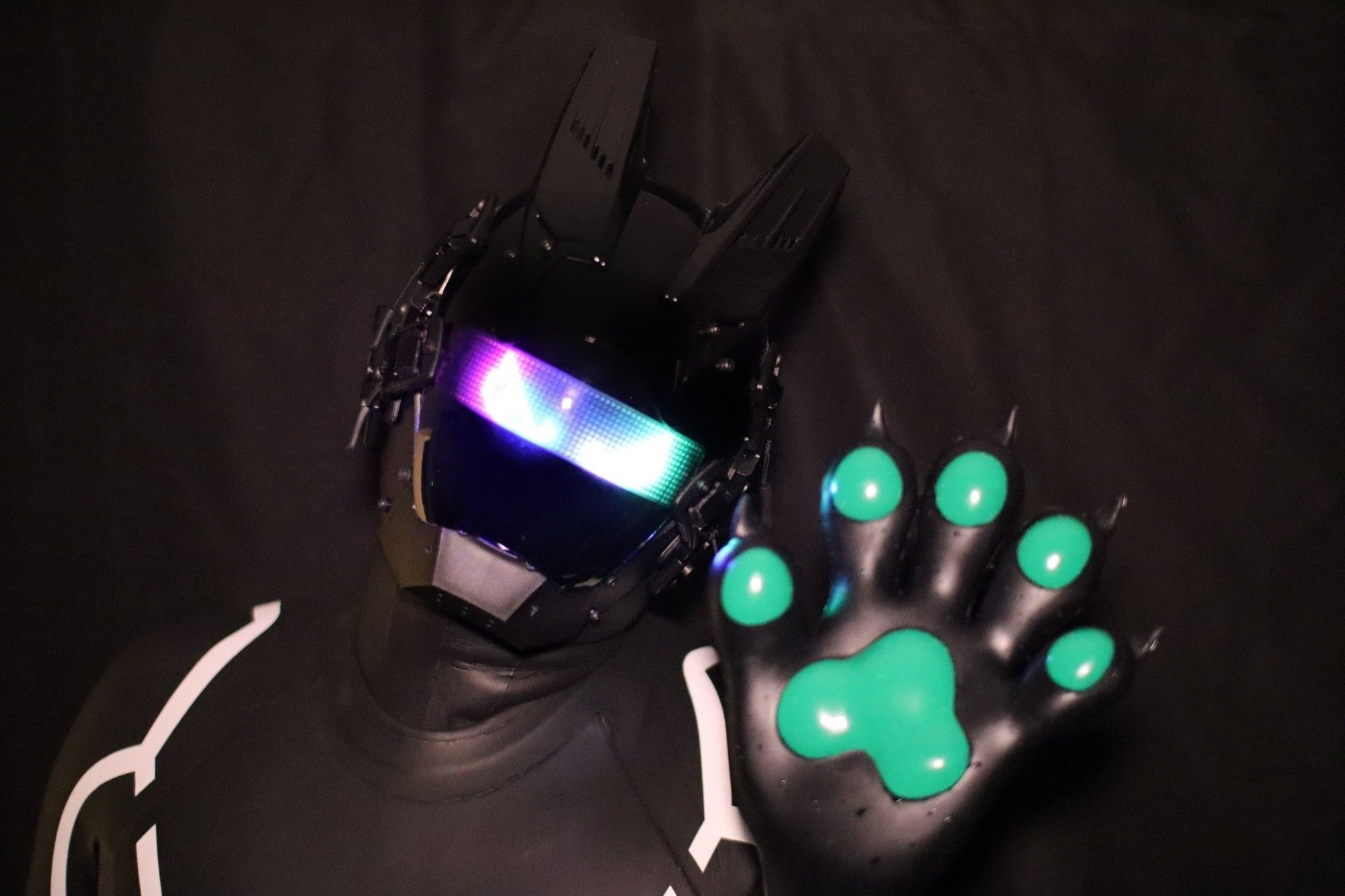 The image shows a person wearing a futuristic, robotic-style helmet with glowing LED lights across the visor, displaying a gradient of purple, blue, and green colors. The helmet has angular, animal-like ears on top. The person is also wearing a black suit with white trim and is holding up a large, black paw with green pads on the palm and fingers, resembling an animal's paw. The background is dark, which makes the glowing elements of the visor and the green pads on the paw stand out vividly.