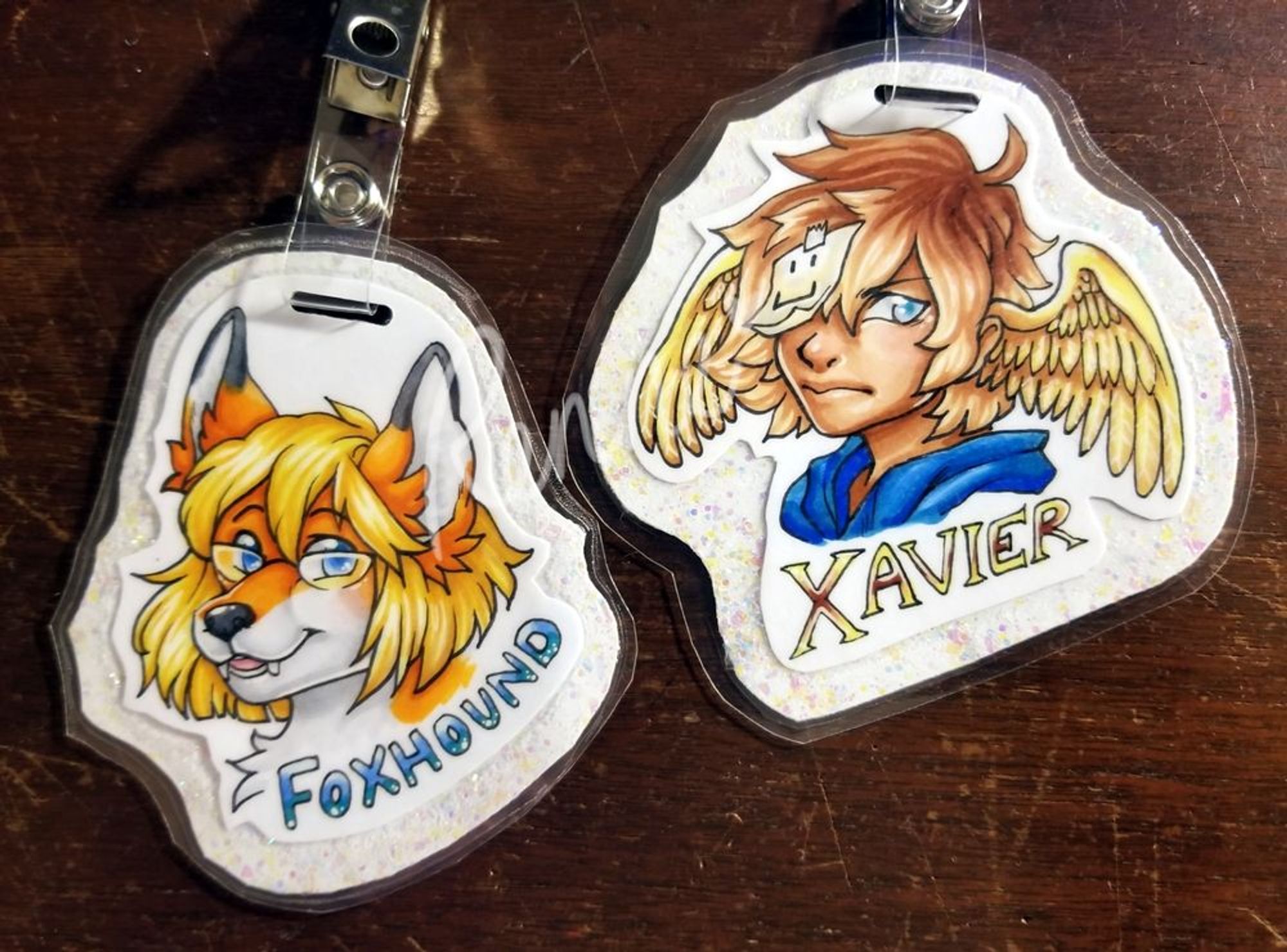 two laminated portraits - one of an anthropomorphic fox, and one of a human boy with wings in his hair. both have clips to be worn as badges.