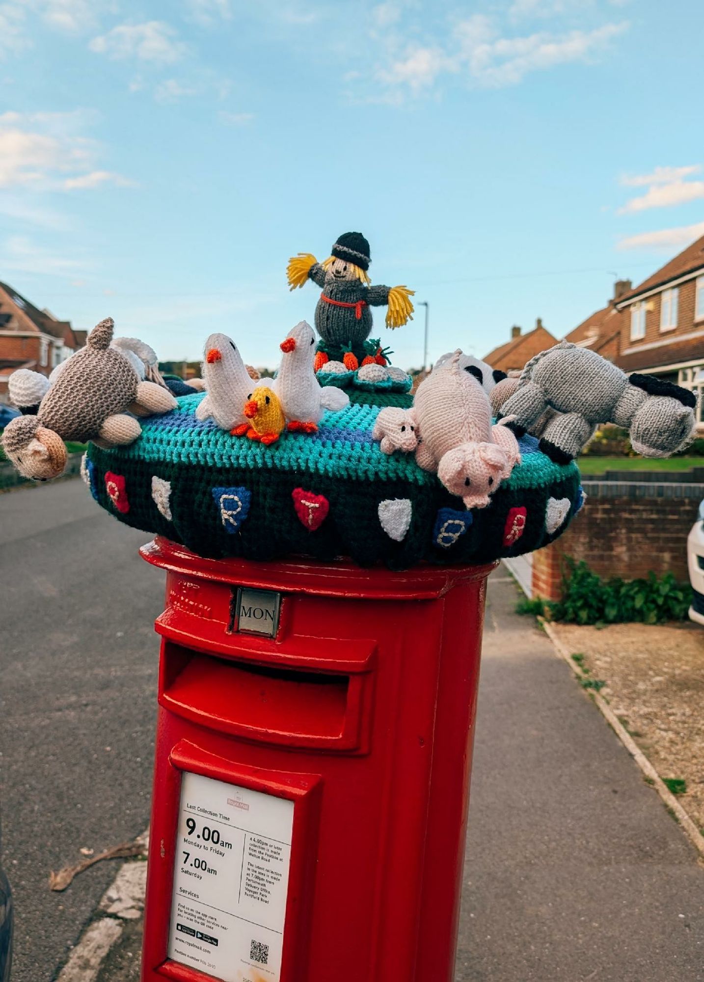 A red postbox on the pavement under a pale blue sky. The postbox has a crocheted topper on it with a scarecrow in the centre and animals around the room peering out and down around the postbox.